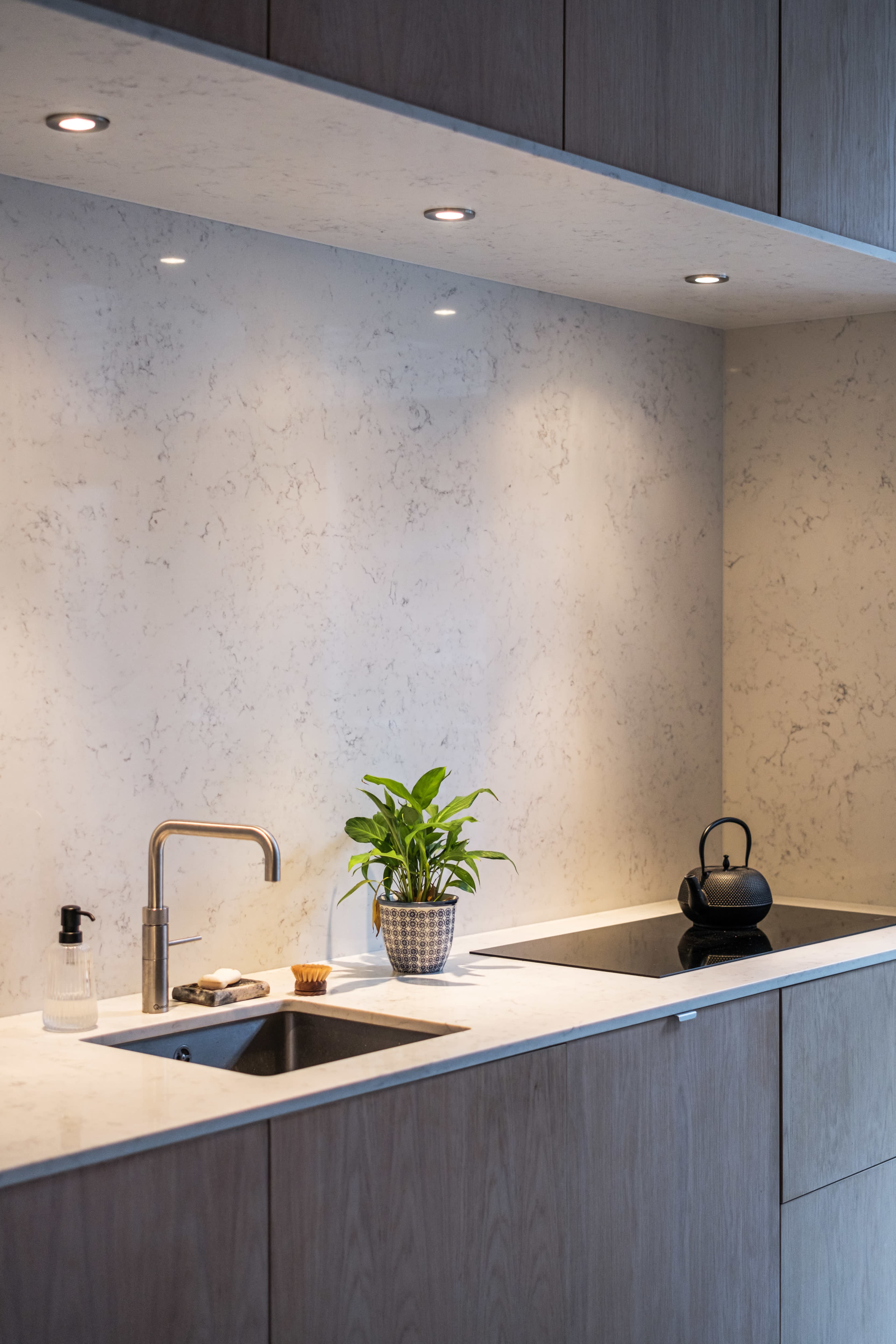 The image shows a modern kitchen with a marble backsplash, a stainless steel sink, a small potted plant, and a black kettle on a countertop.
