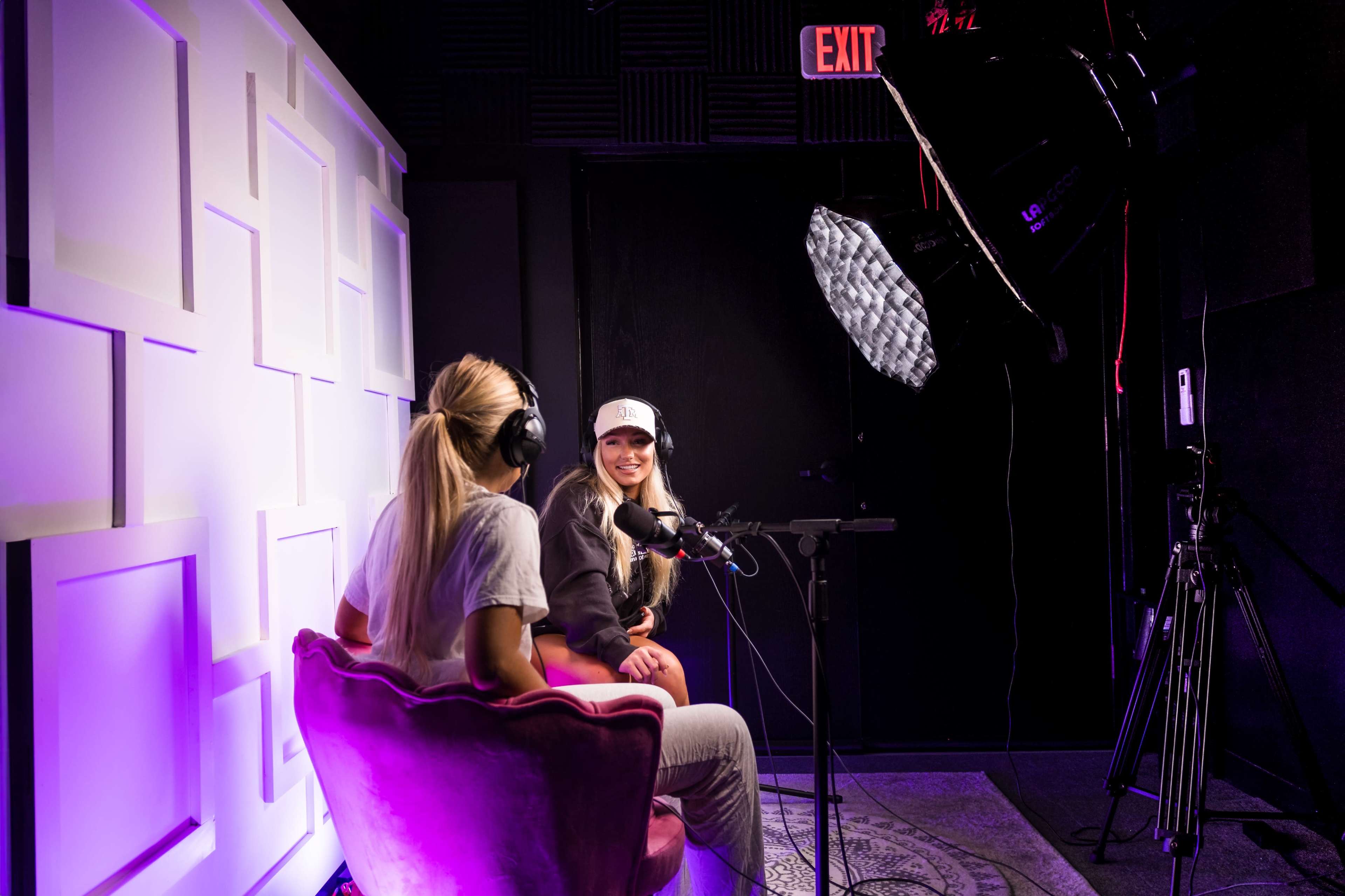 Two women sit in a studio with microphones, engaging in conversation while wearing headphones, in front of a lit background.