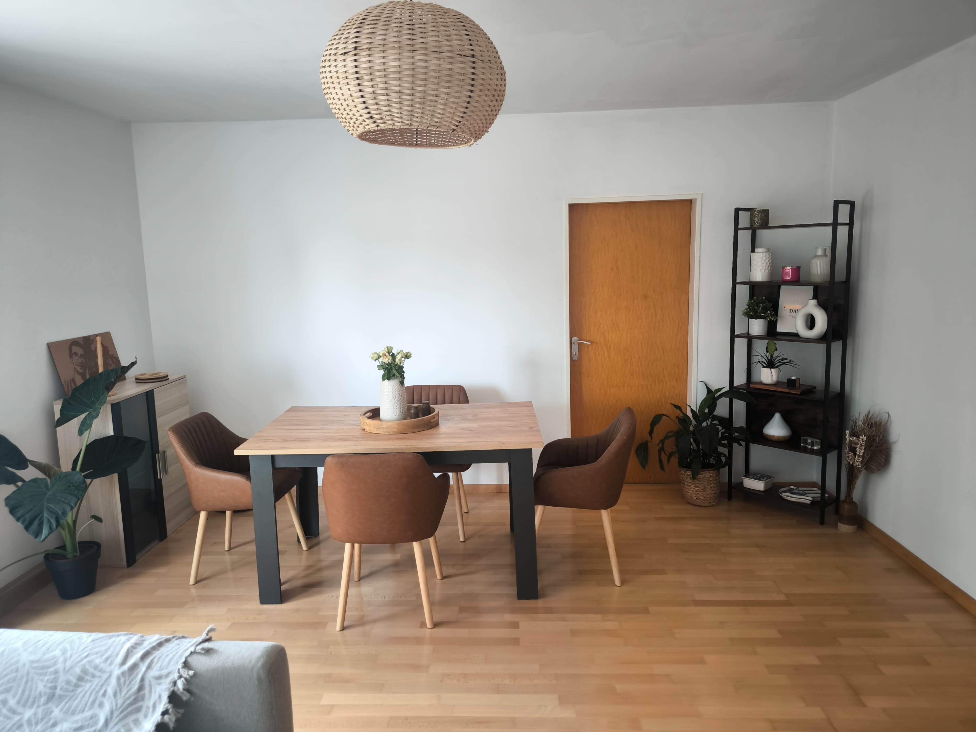 A dining area features a wooden table surrounded by four brown chairs, with a bookshelf and potted plants nearby.