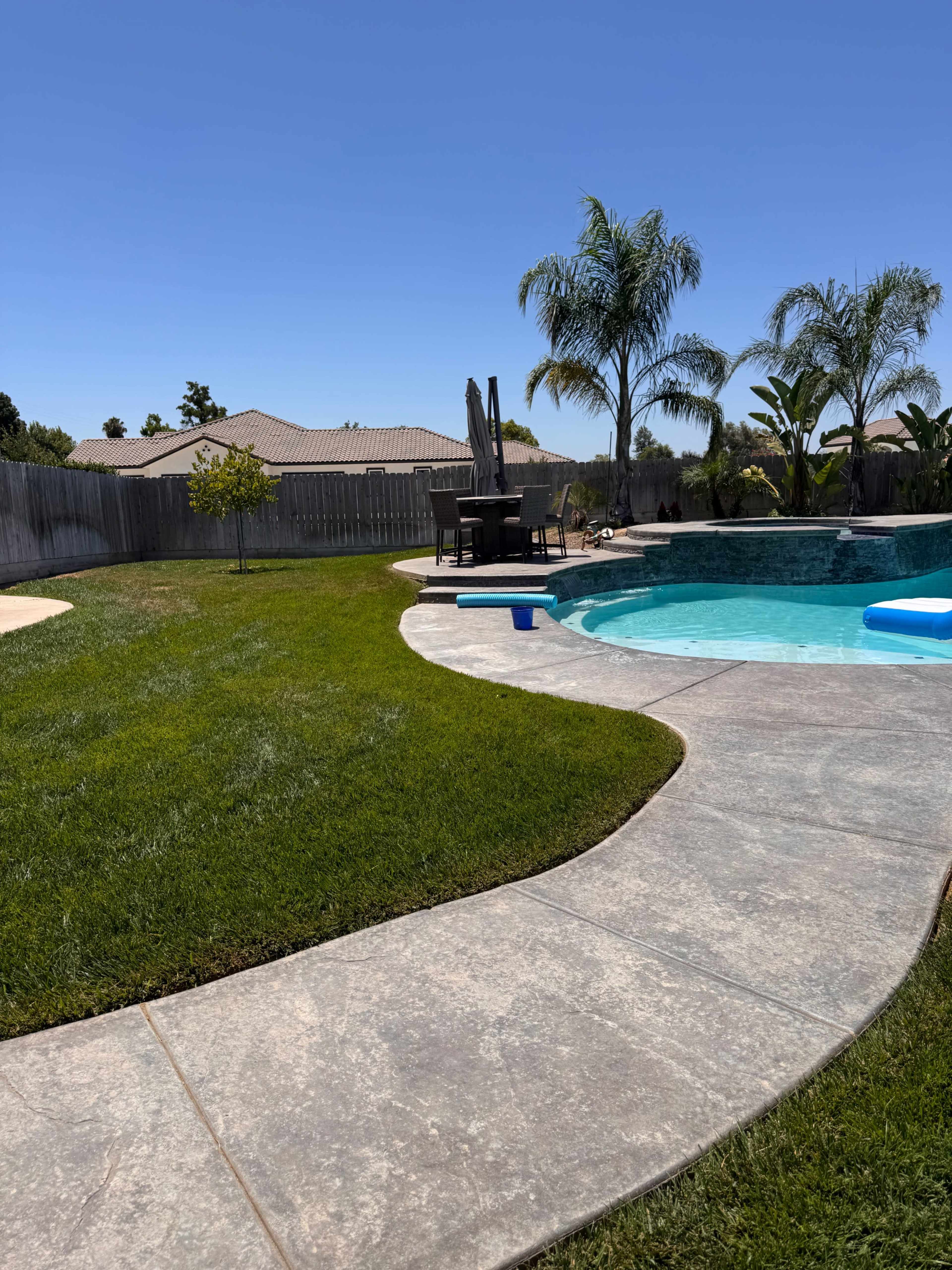 A backyard features a blue swimming pool, a stone pathway, and palm trees, with a house visible in the background.
