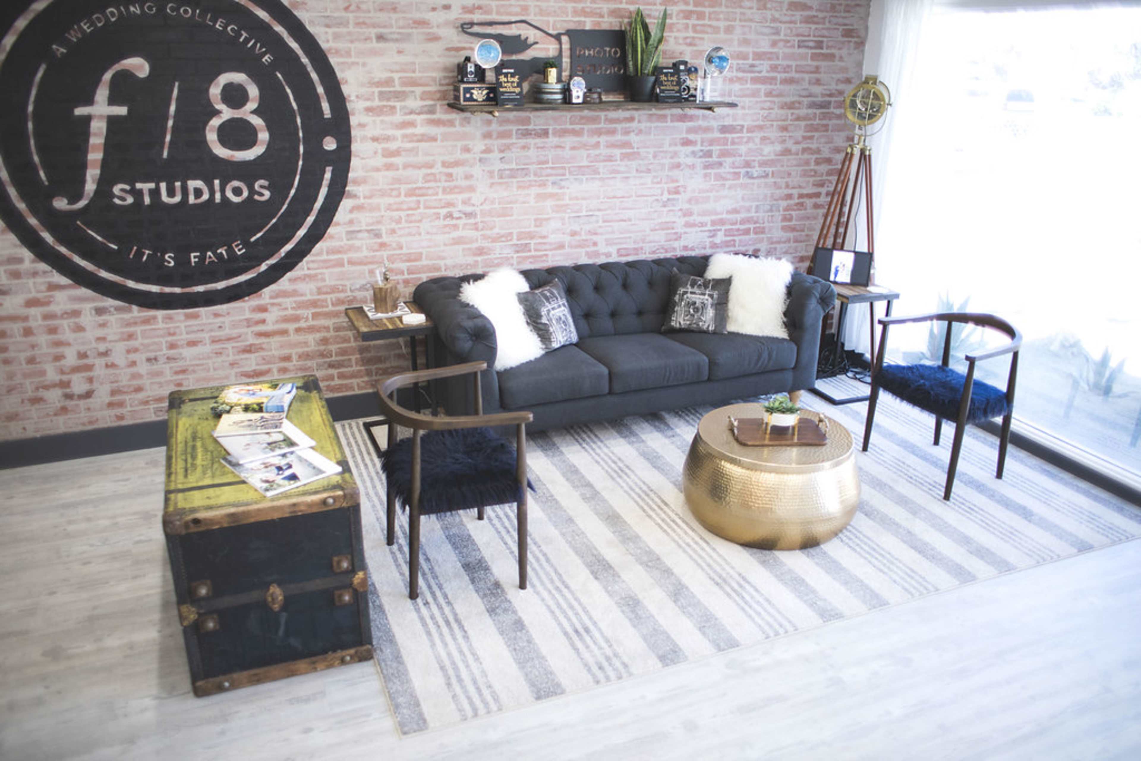 A modern studio interior featuring a dark sofa with pillows, a round gold coffee table, two black chairs, and a chest at the side, all arranged against a brick wall with a circular logo.