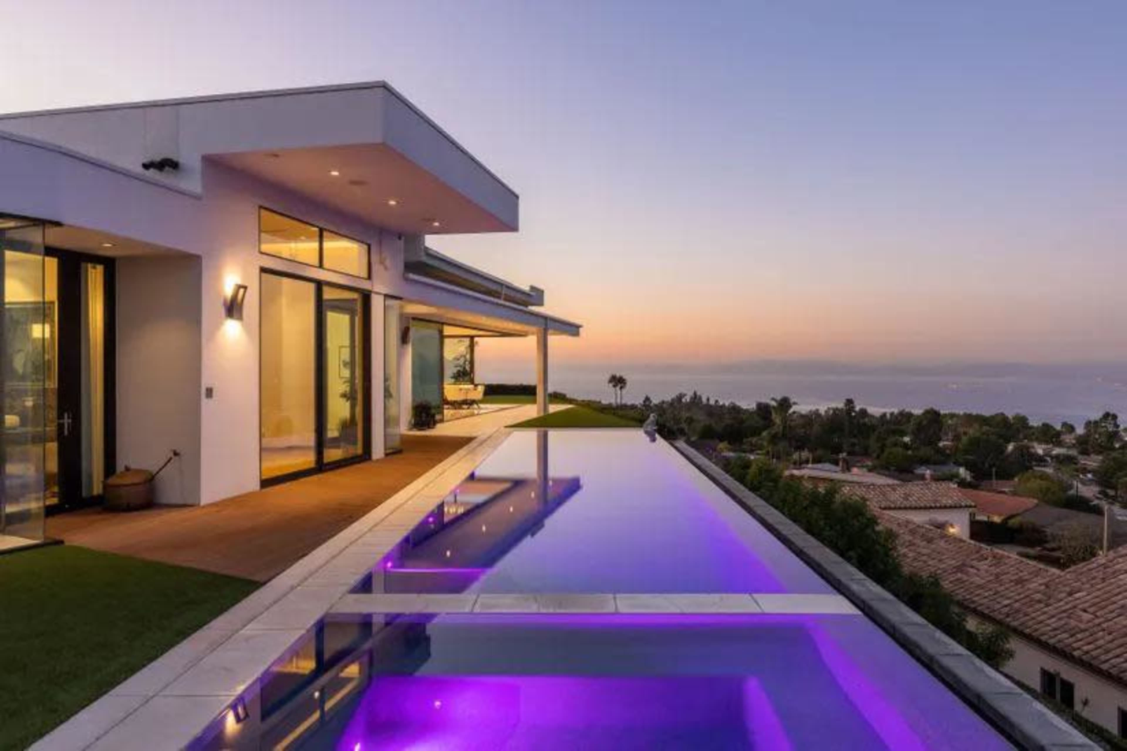A modern house with large glass windows overlooks a pool illuminated with purple lights at sunset.