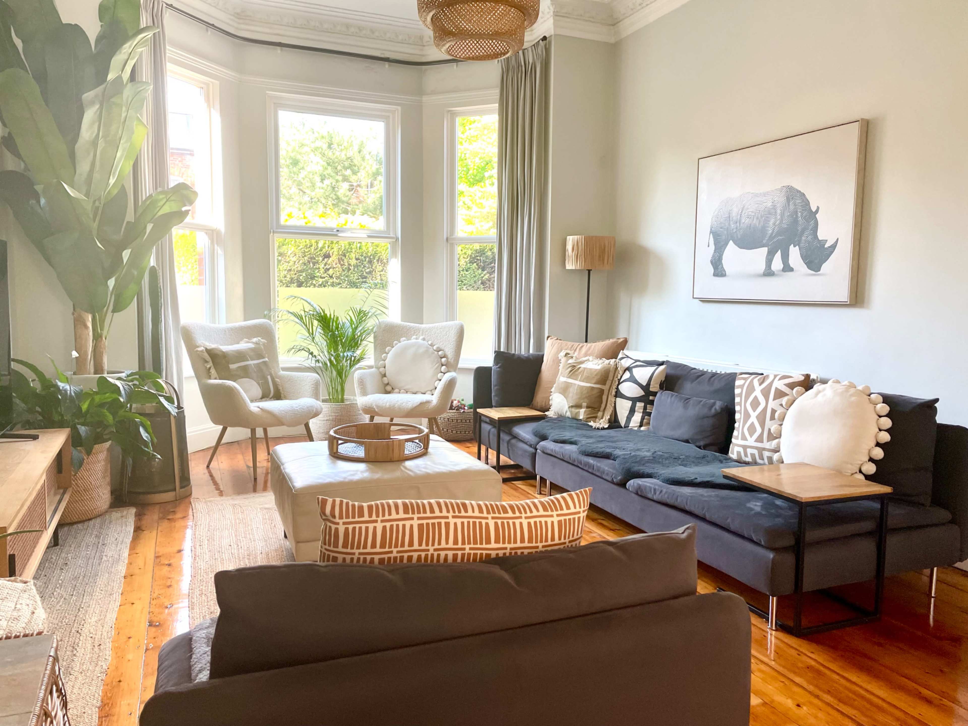 A bright living room features a mix of seating arrangements, potted plants, and a large rhino artwork on the wall.