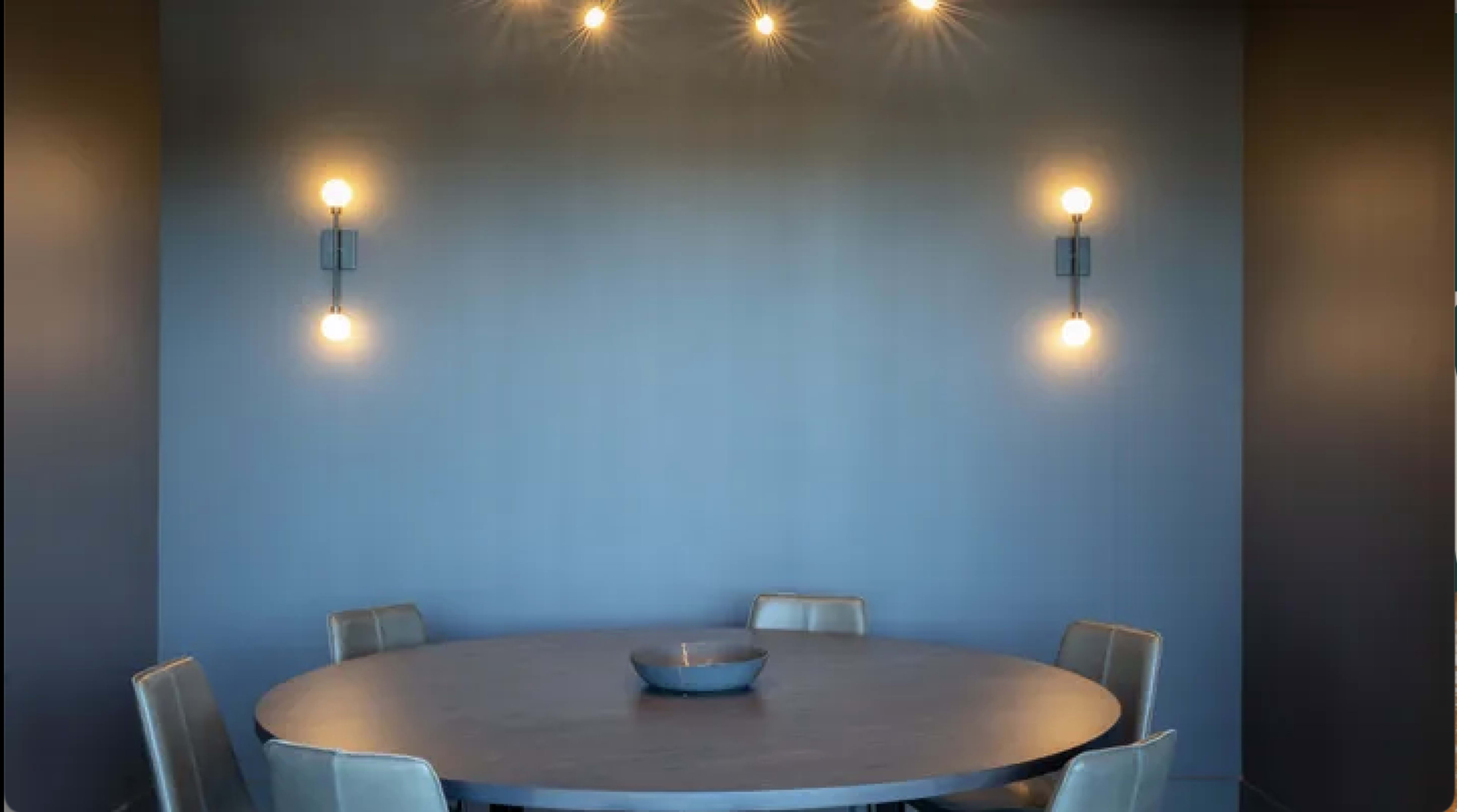 A round dining table with five chairs is set in a room featuring a textured blue wall and overhead lighting.
