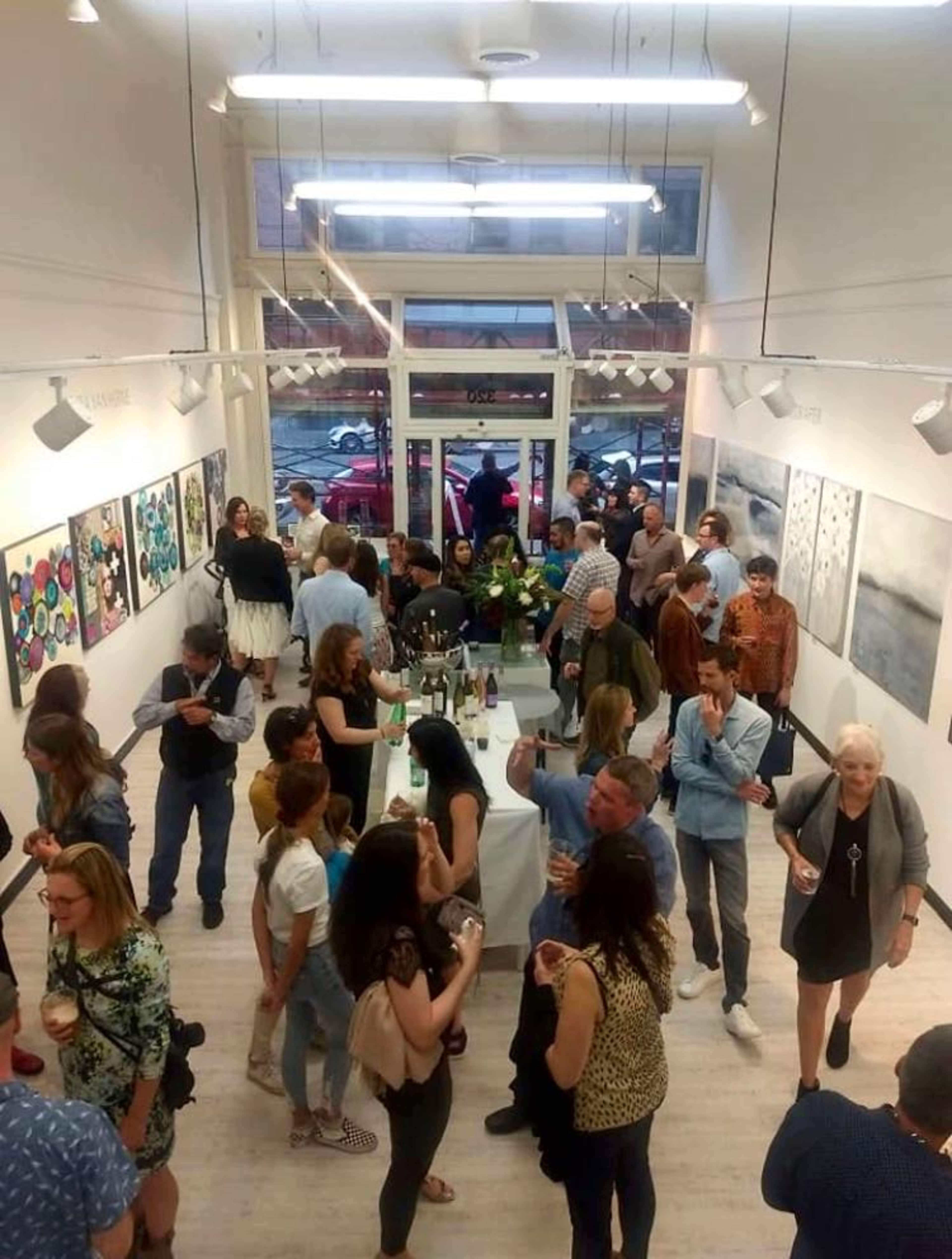 A large group of people is mingling at an art gallery opening, surrounded by colorful artworks and refreshment tables.