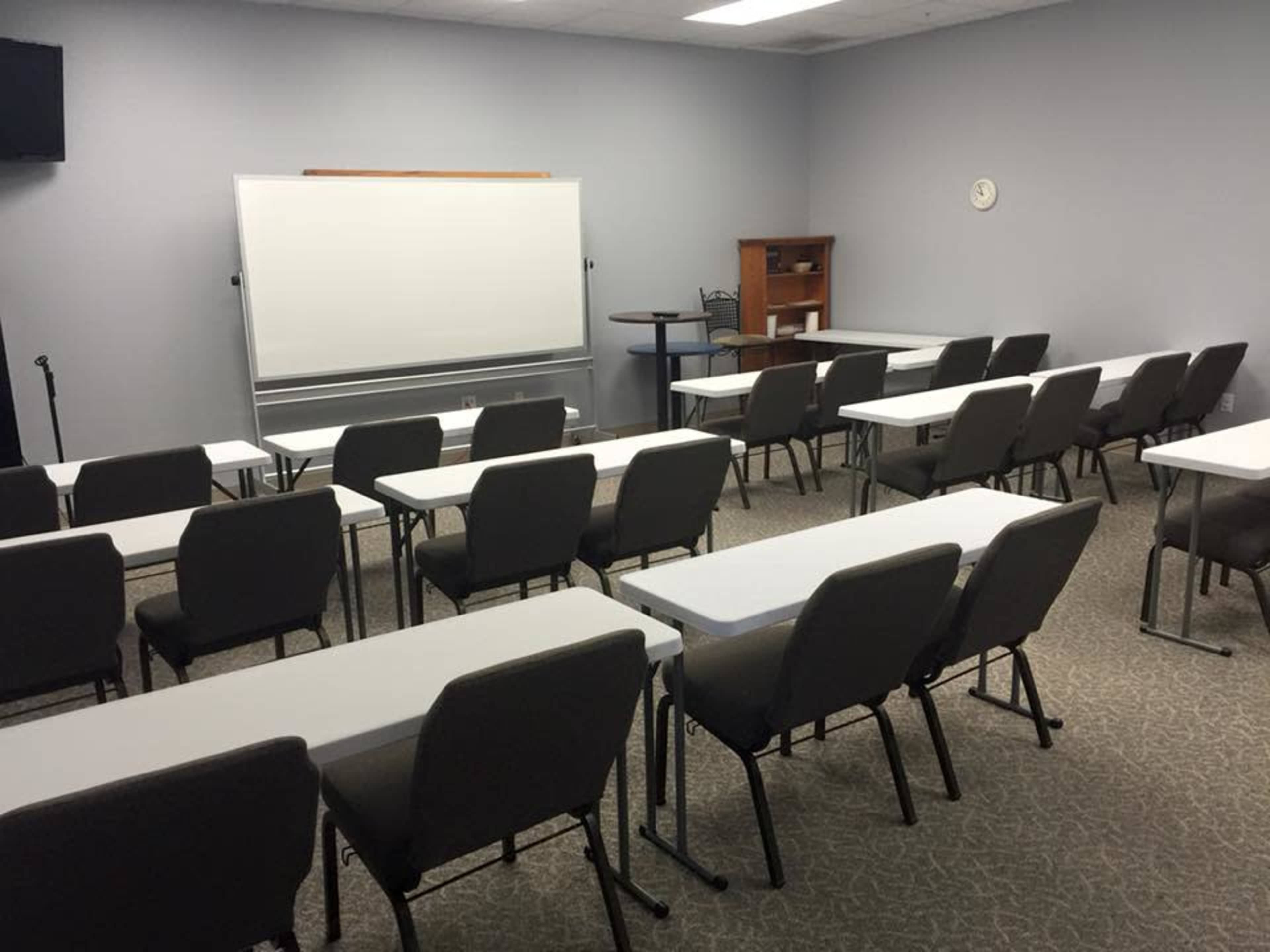 The image shows a classroom setup with rows of tables and chairs, a whiteboard at the front, and a small bookshelf in the corner.