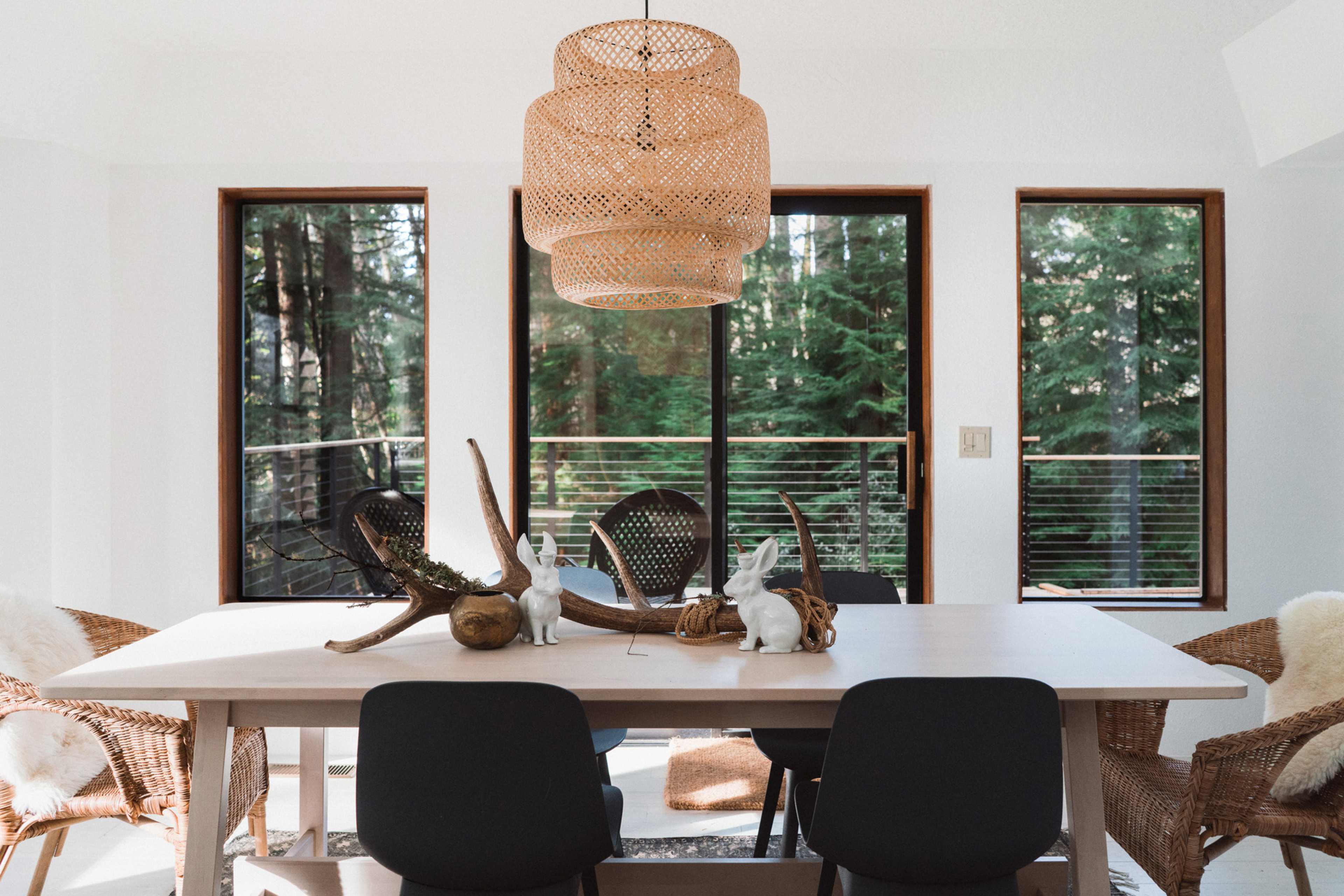 A dining table is set in a bright room with large windows overlooking a forest, adorned with decorative items and surrounded by woven chairs.