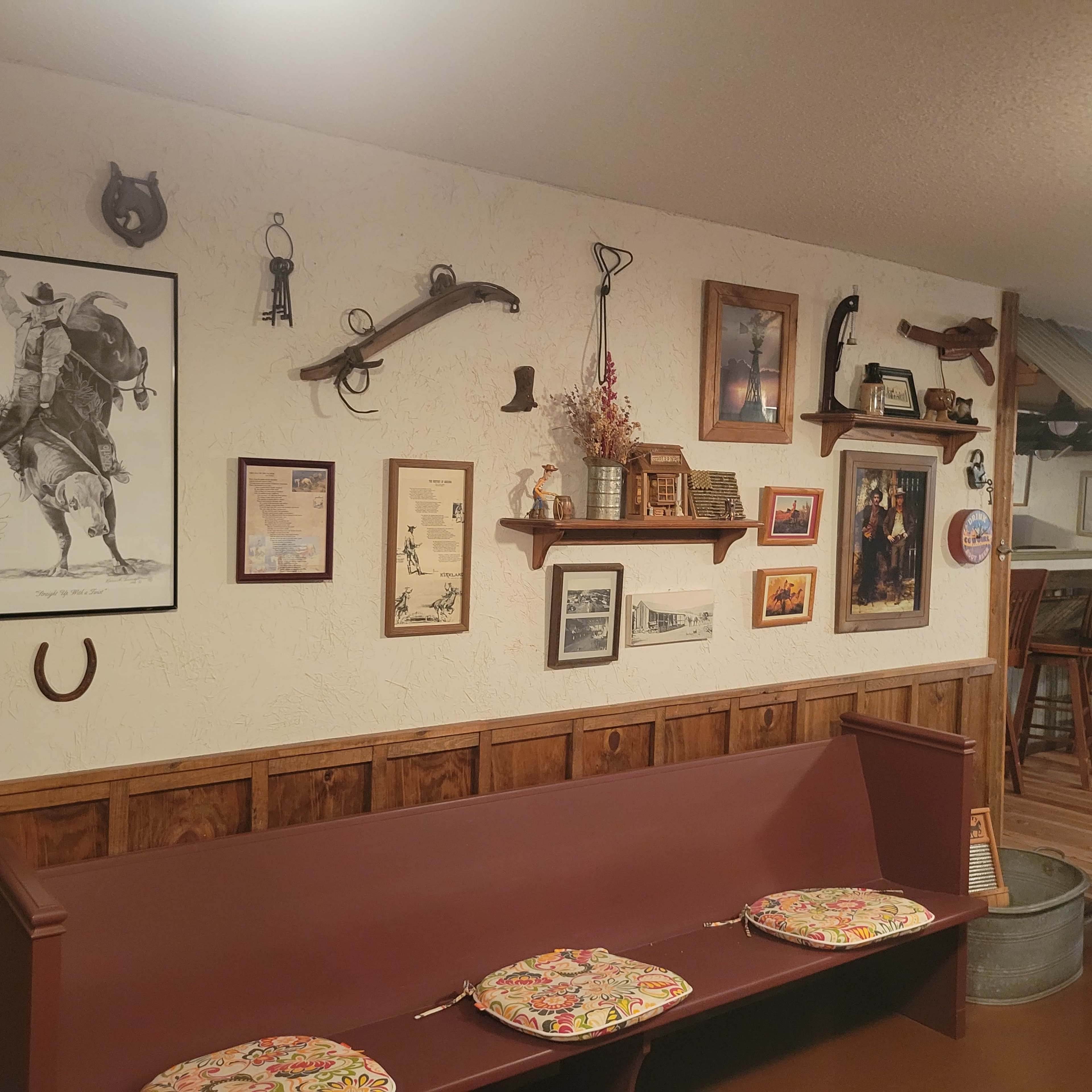 The image shows a rustic interior wall adorned with Western-themed art, vintage tools, shelves of memorabilia, and a wooden bench with patterned cushions.