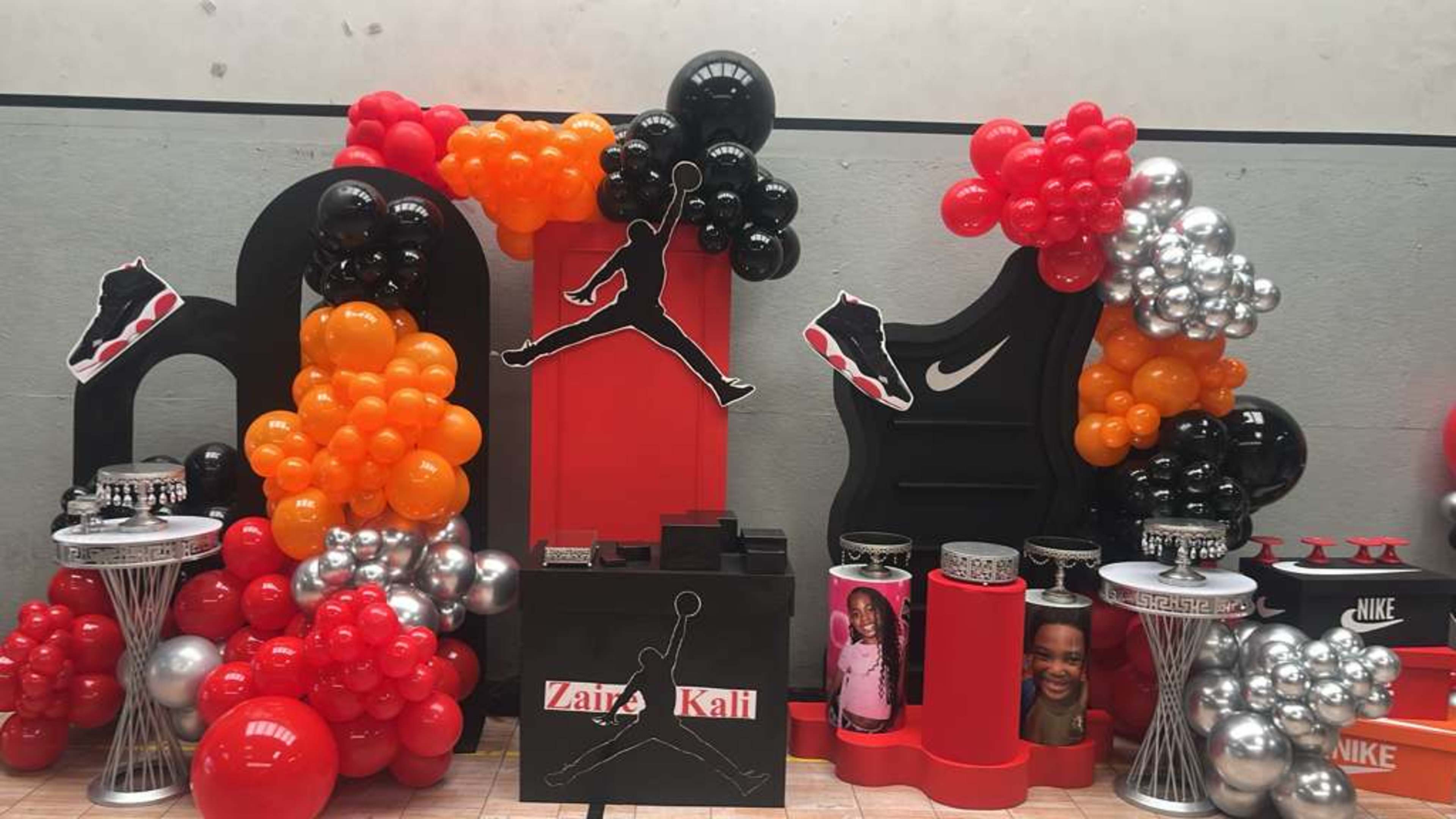 The image depicts a festive display featuring black, orange, and silver balloons arranged around a central black and red Jordan logo, alongside tables with various themed decorations.