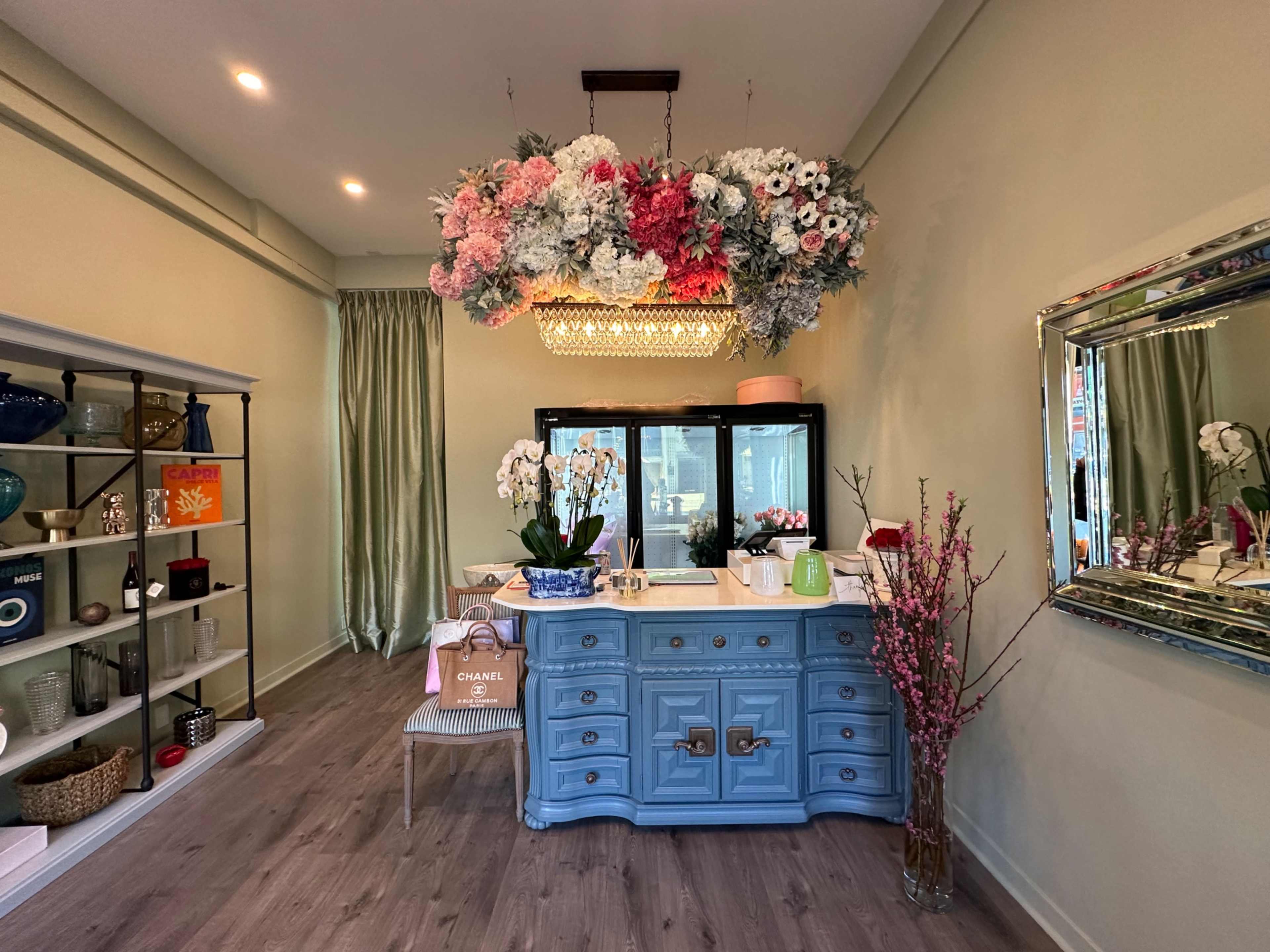 A light blue dresser with decorative flowers overhead stands in a softly lit room featuring a mirror and shelves filled with various items.