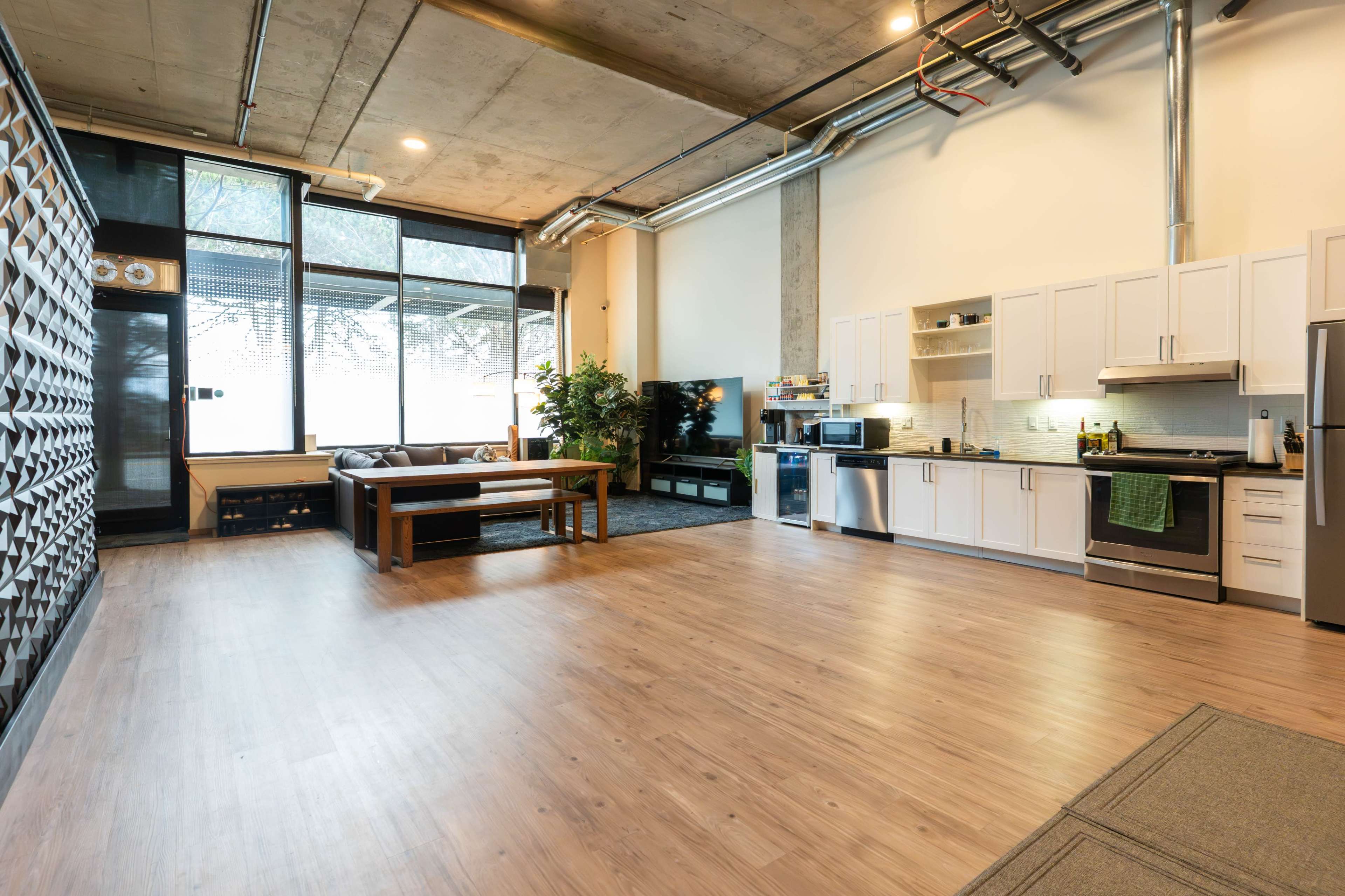 The image shows a spacious, modern kitchen and living area featuring a large window, a couch, and a kitchen with white cabinets and stainless steel appliances.