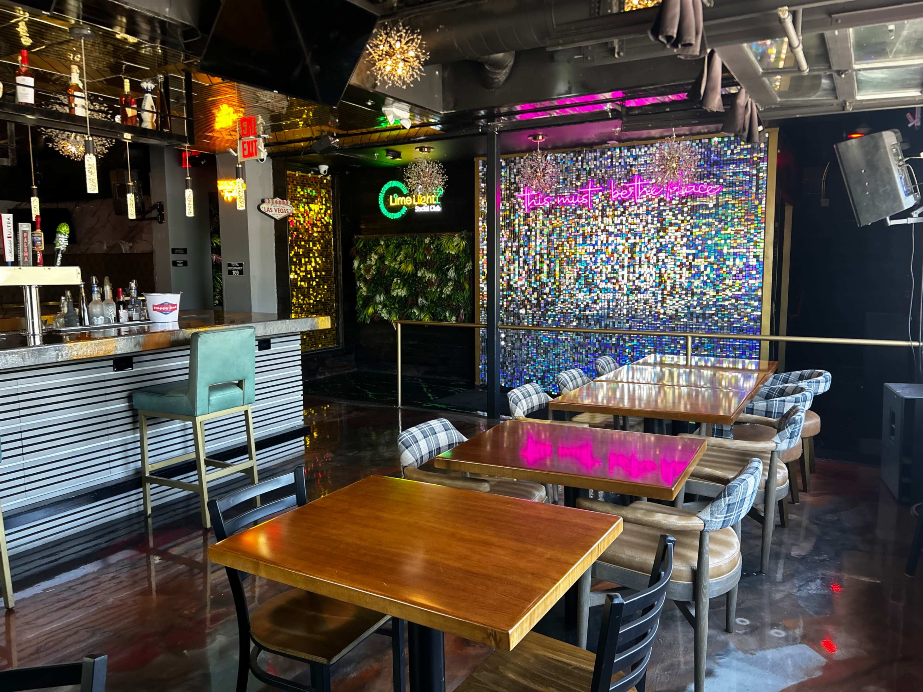 The interior of a modern bar features colorful lighting, a decorative wall made of small tiles, and wooden tables with black chairs.