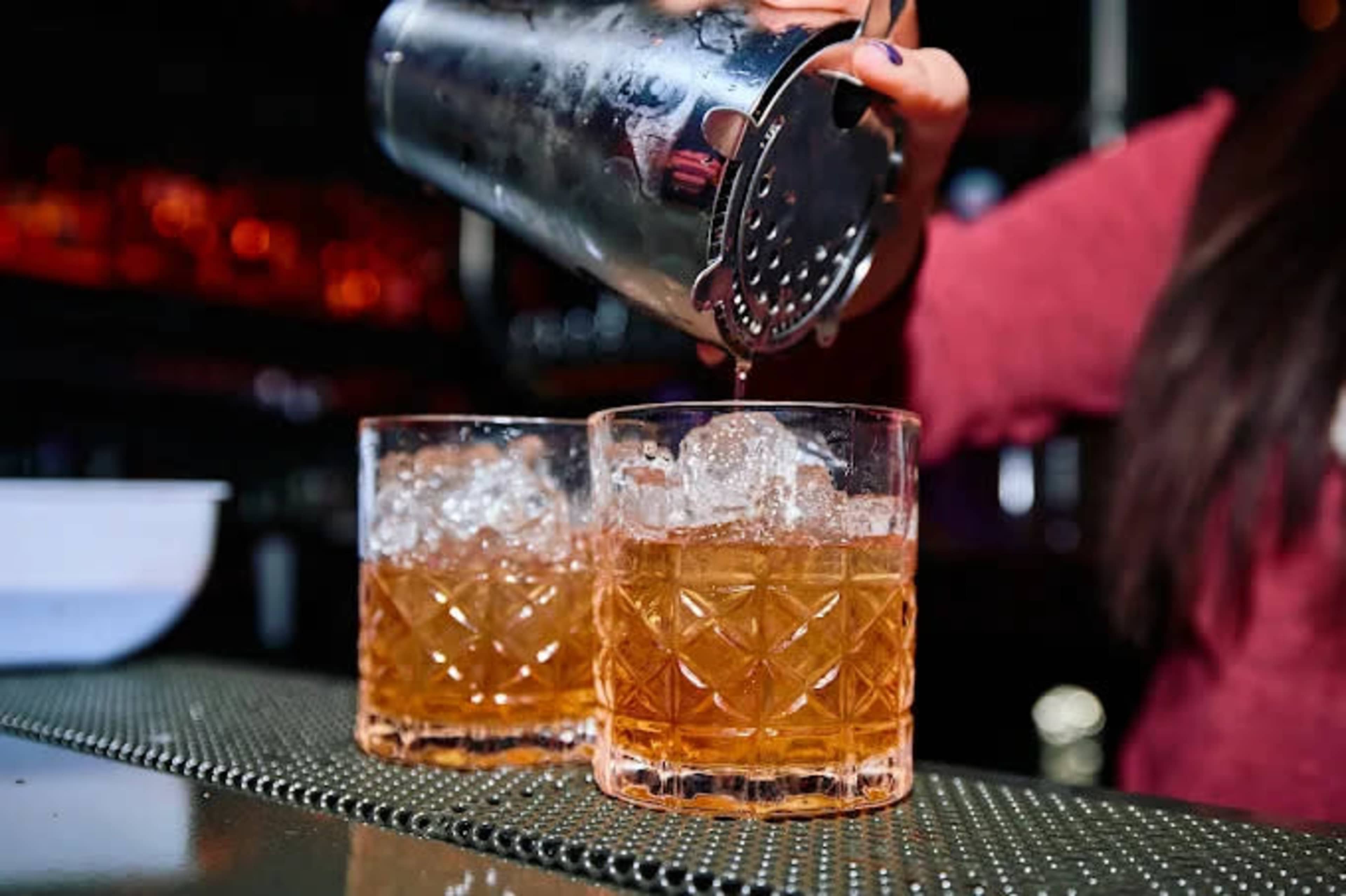 A bartender pours a cocktail from a shaker into two glasses filled with ice.