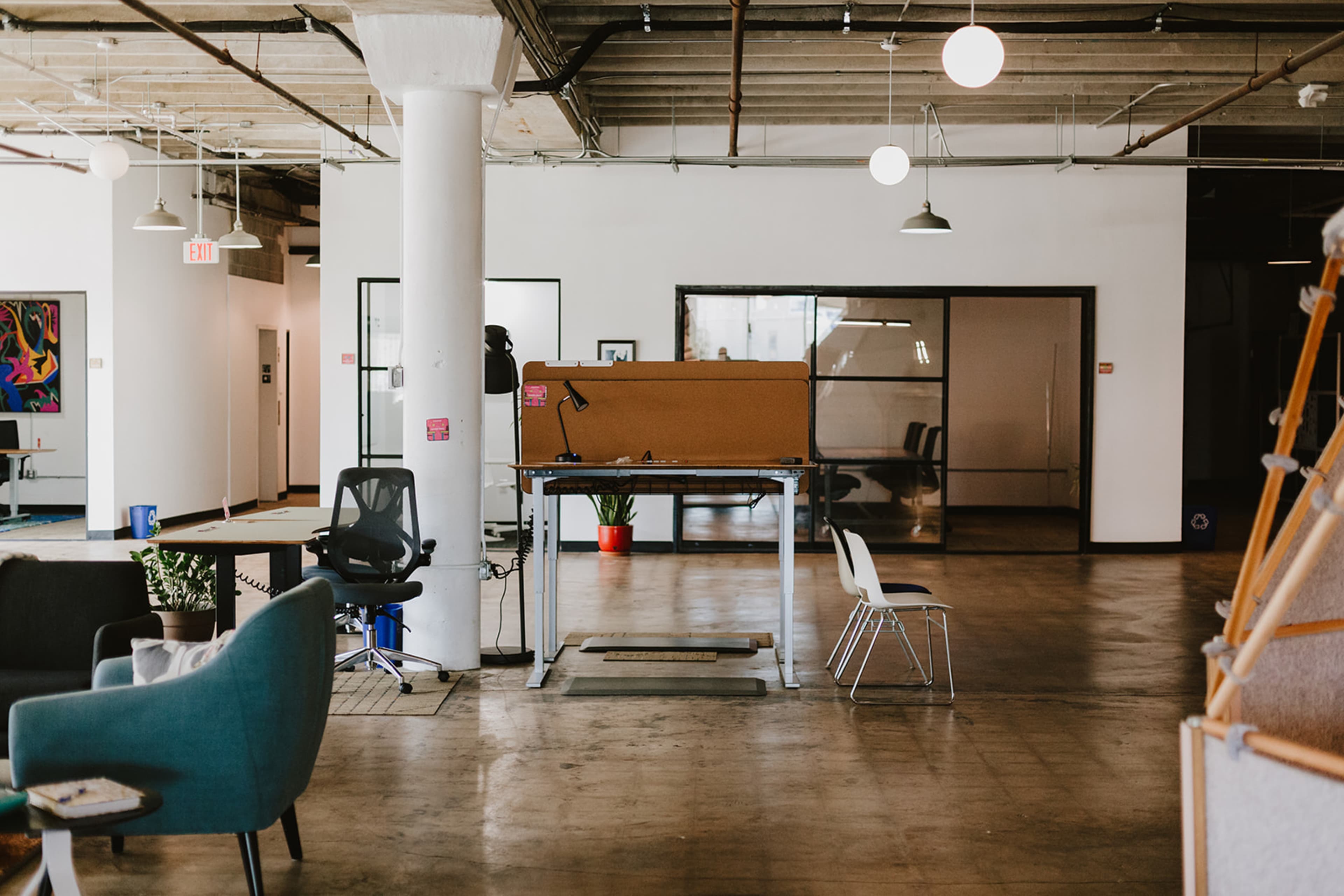 The image shows a spacious office interior with a standing desk, chairs, and various plants, featuring a minimalist design and large windows.