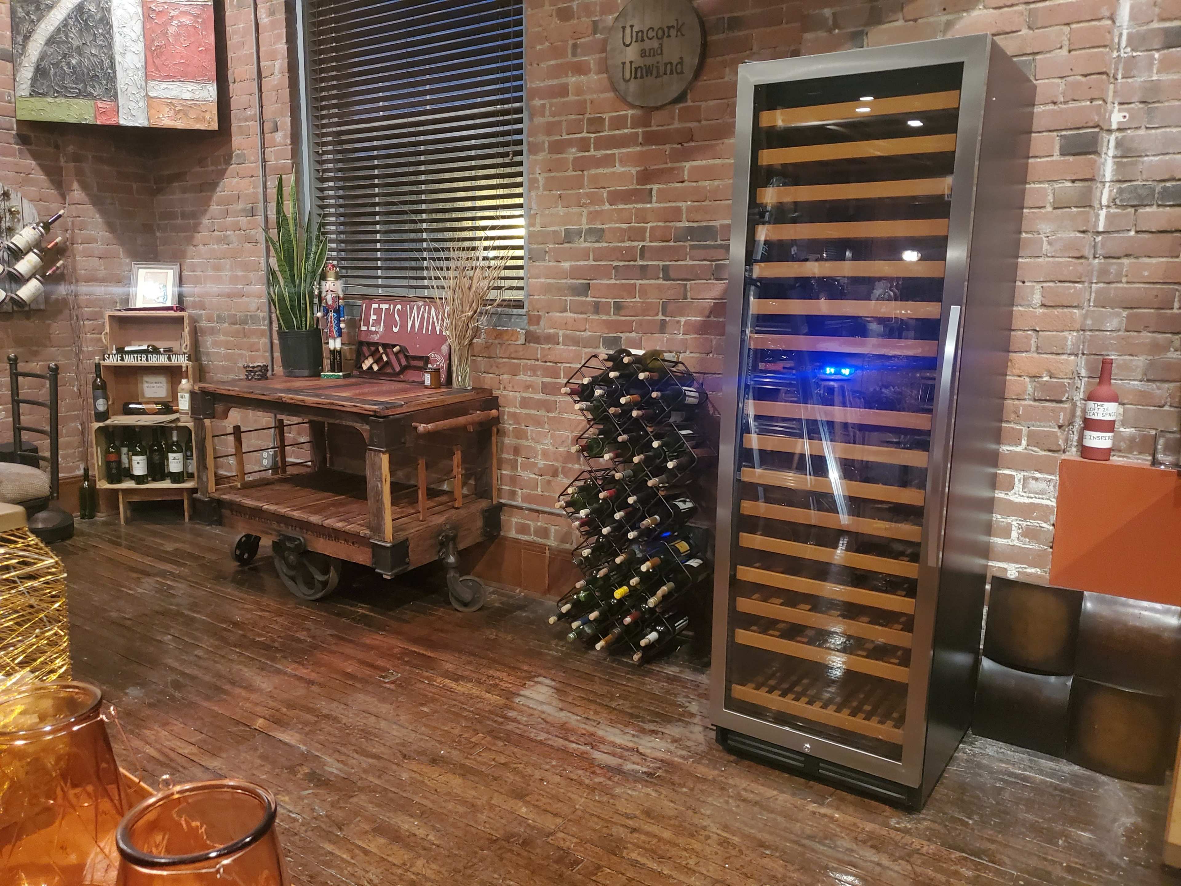 A modern wine refrigerator beside a wooden cart and a wall-mounted wine rack in an industrial-style room with exposed brick.