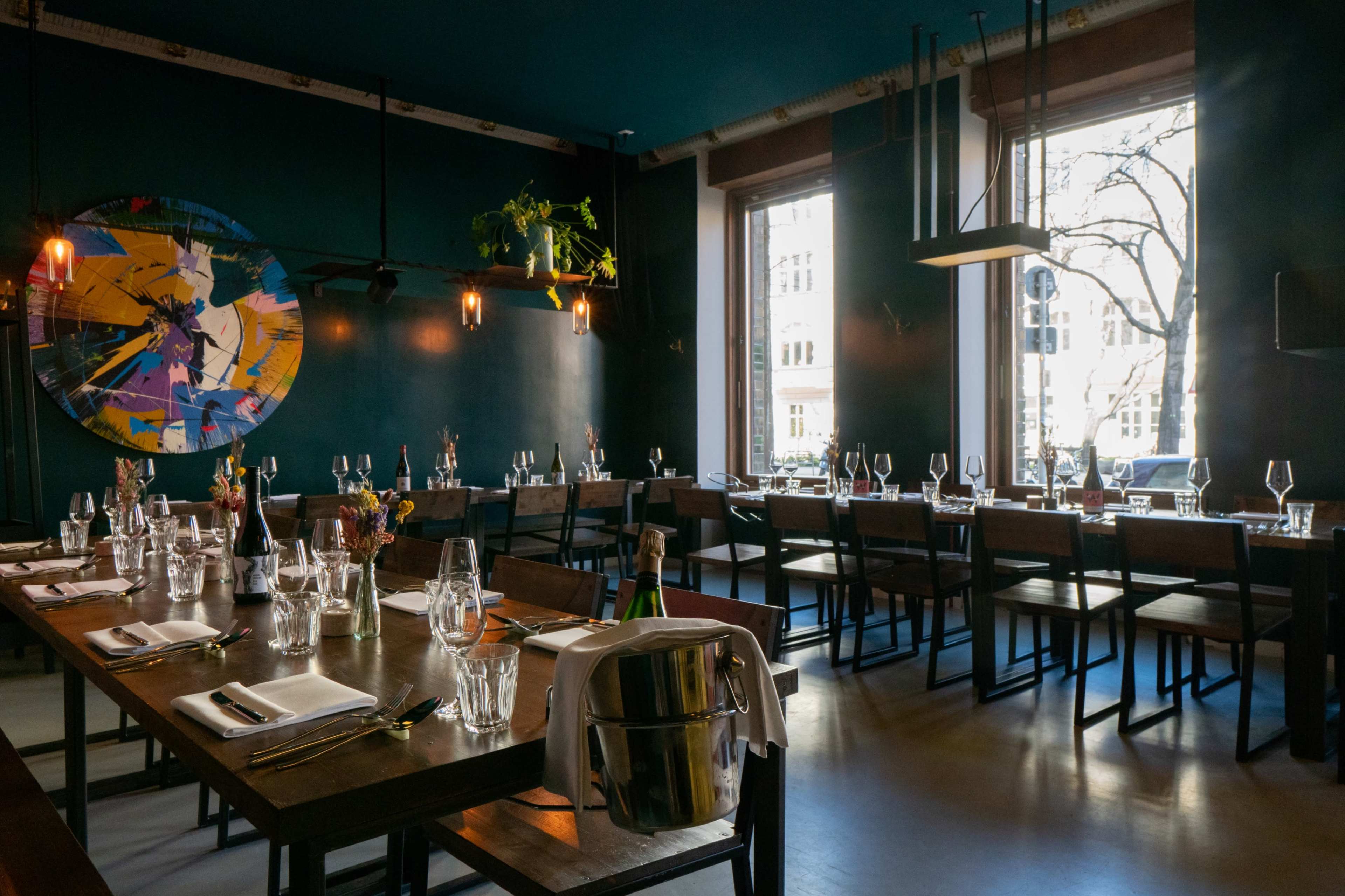 The image shows a modern restaurant interior with a long wooden table set for dining, surrounded by dark blue walls and large windows allowing natural light to enter.