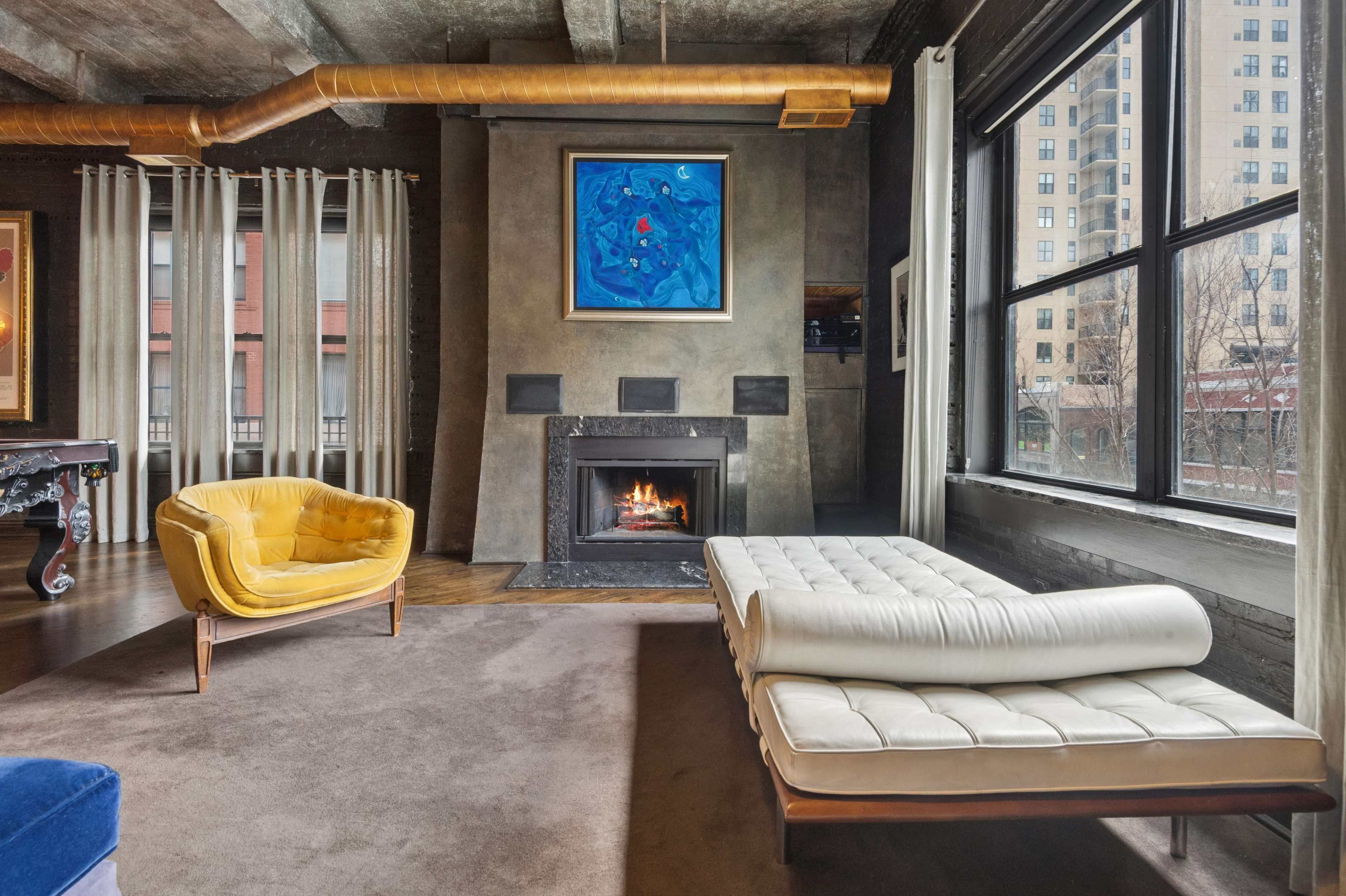 The image shows a modern living room with a yellow armchair, a white sofa, a fireplace, and large windows overlooking a cityscape.