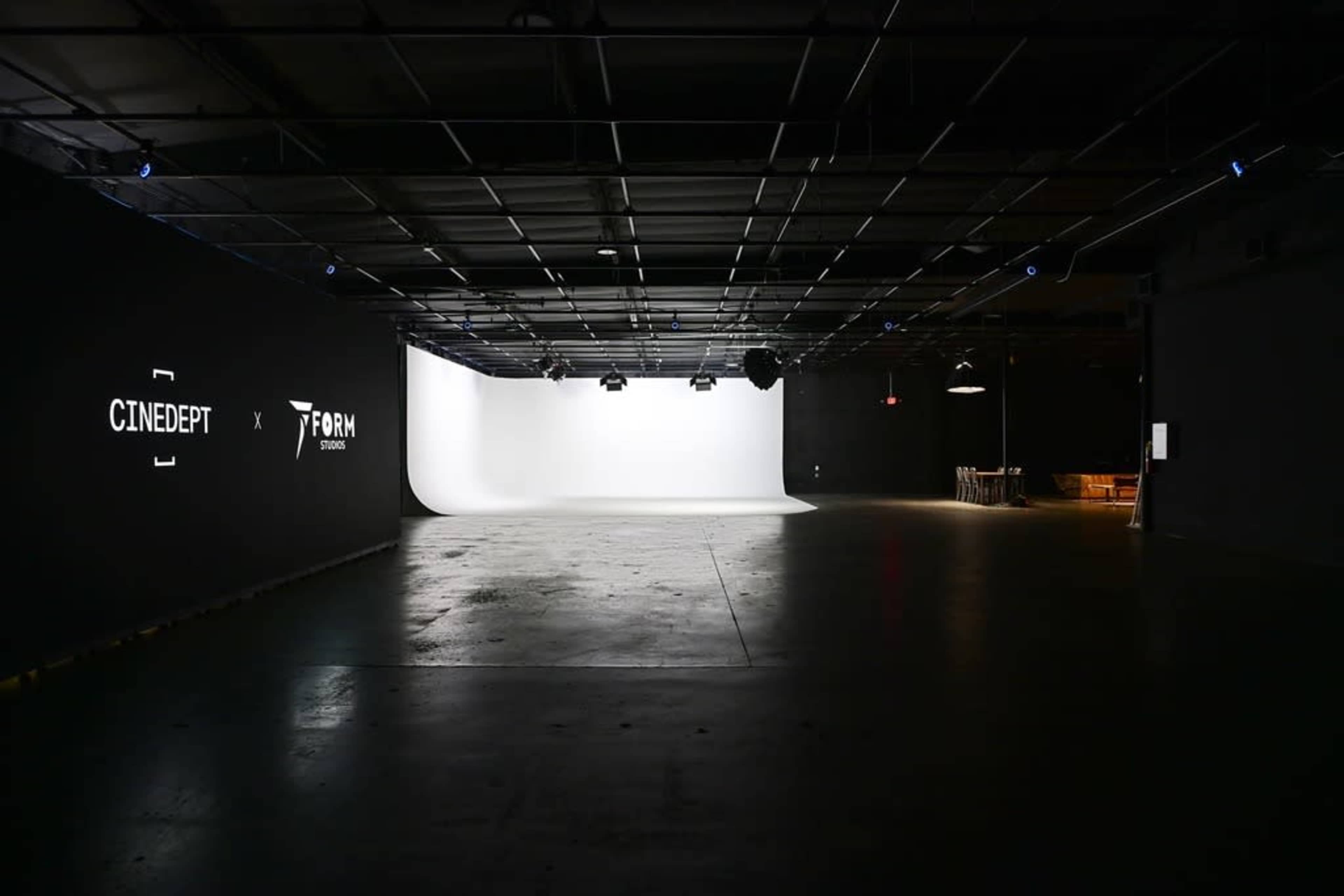 The image shows a spacious studio interior with a bright white backdrop on one side and dark walls featuring logos on the left side.