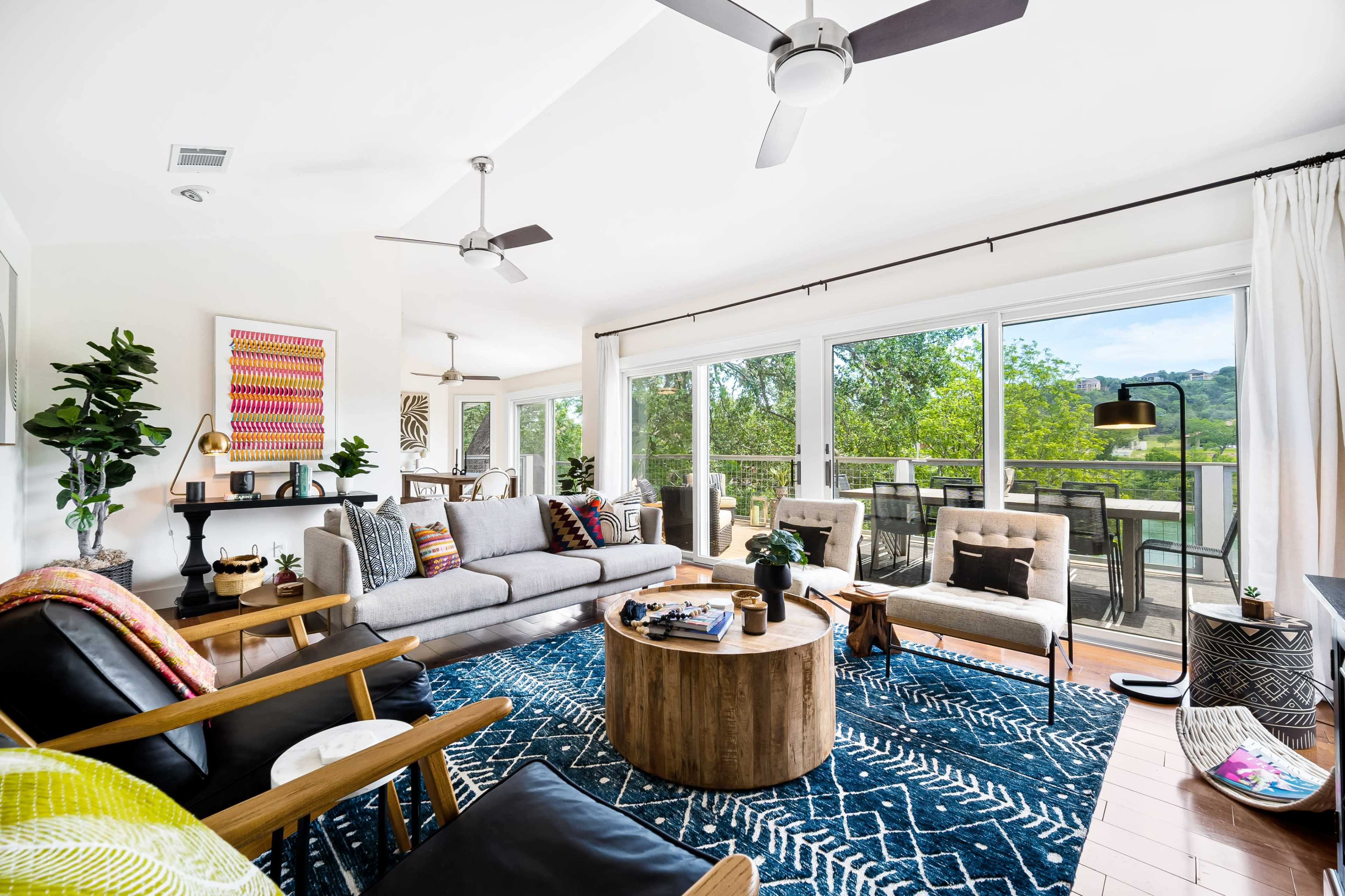 The living room features a gray sofa, wooden coffee table, and large windows overlooking a green landscape.