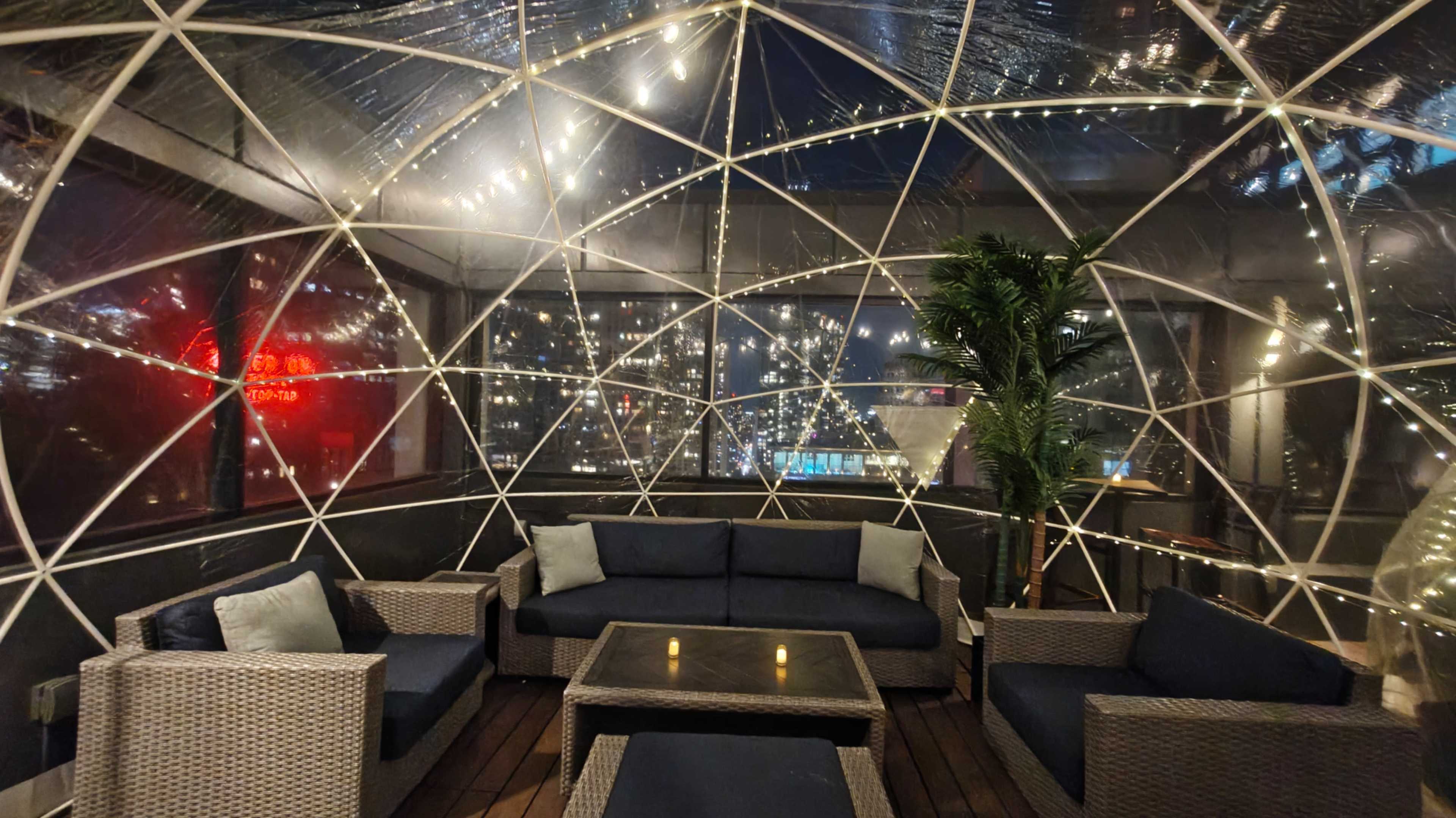The image shows a cozy, dome-shaped seating area with a few couches and a table, surrounded by city lights visible through transparent walls.