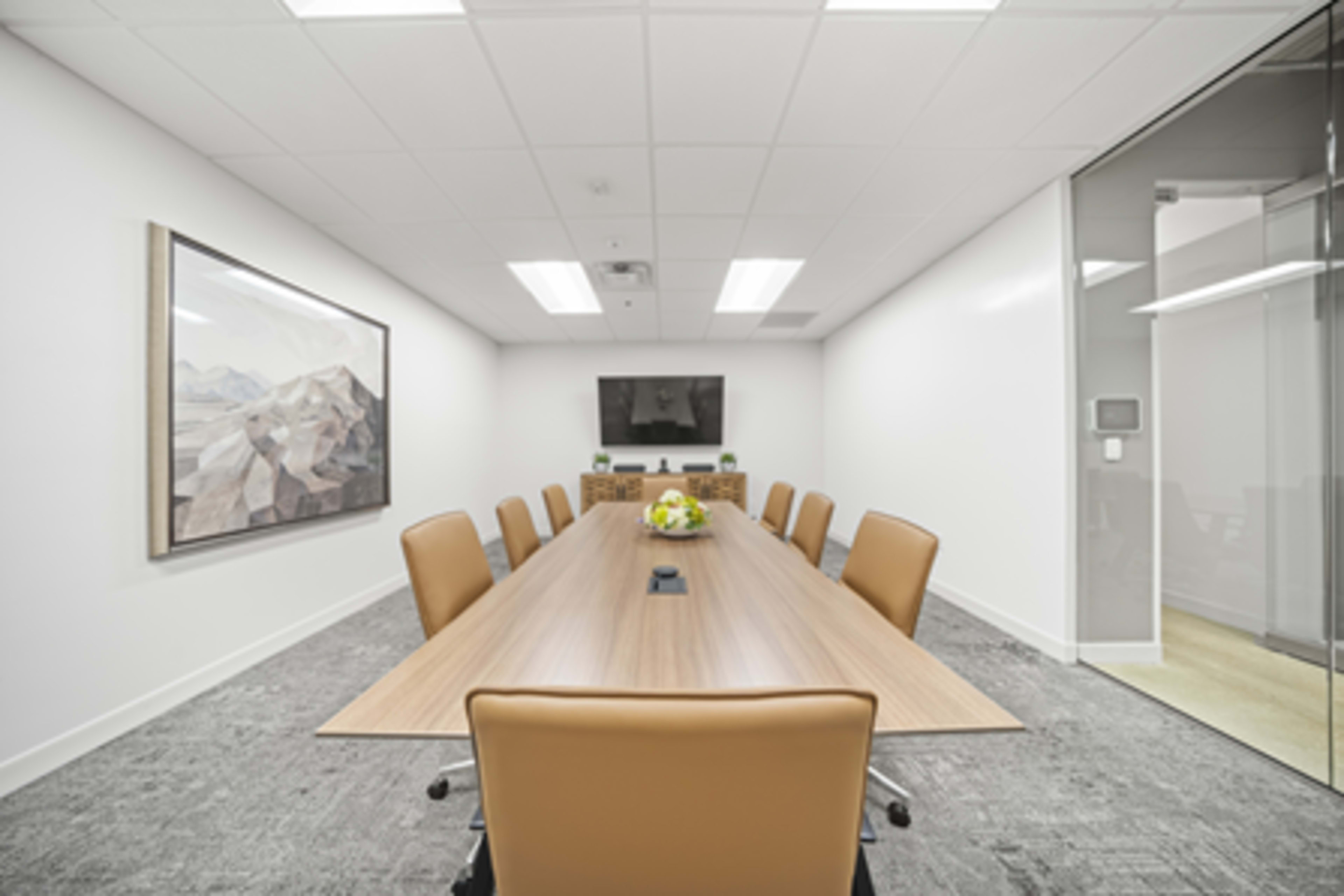 The image shows a modern conference room featuring a long wooden table with eight tan chairs, a television on the wall, and a framed landscape painting.