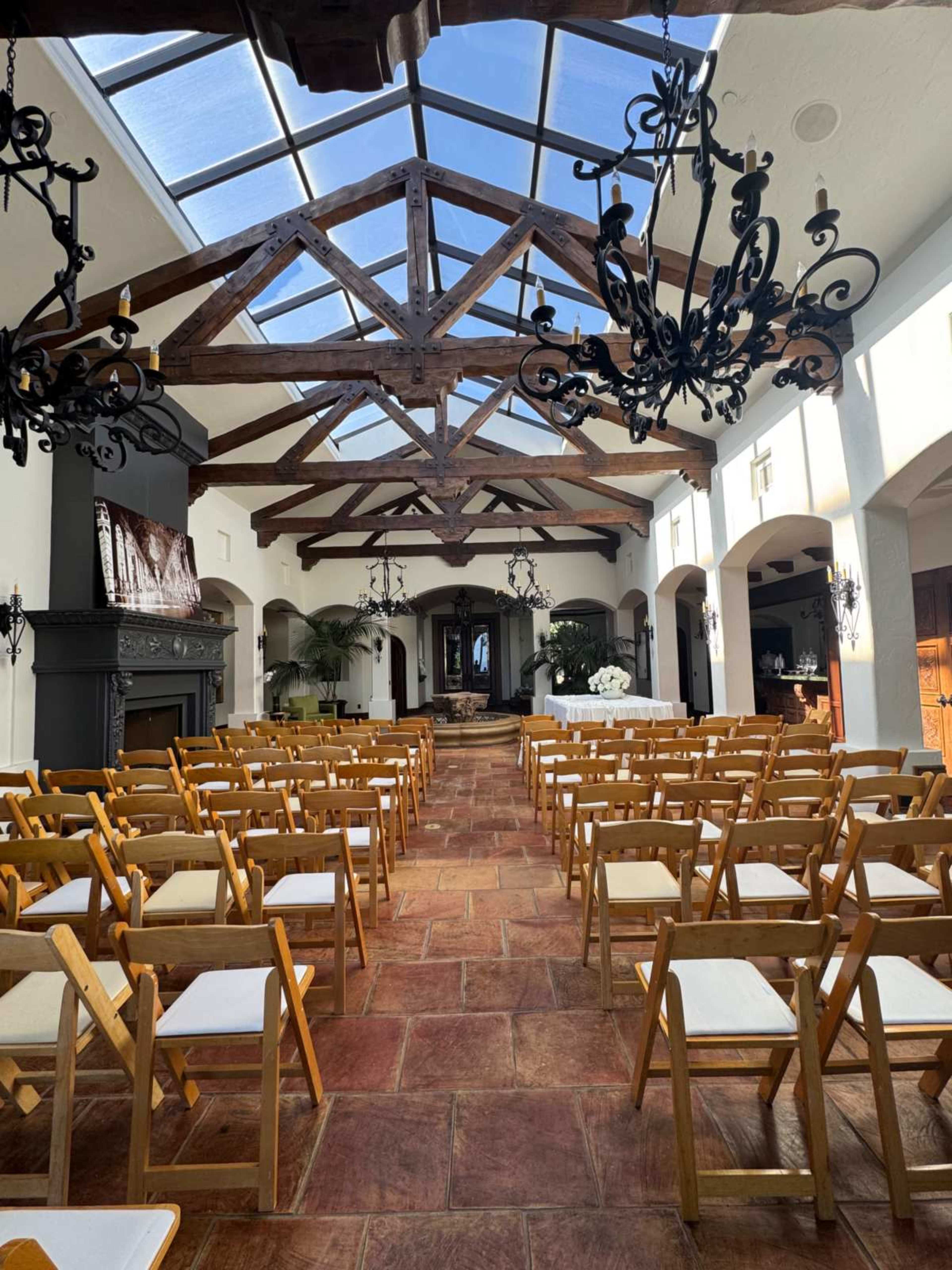A spacious indoor venue features wooden beams, chandeliers, and rows of folding chairs arranged for an event, with a large window allowing natural light to enter.