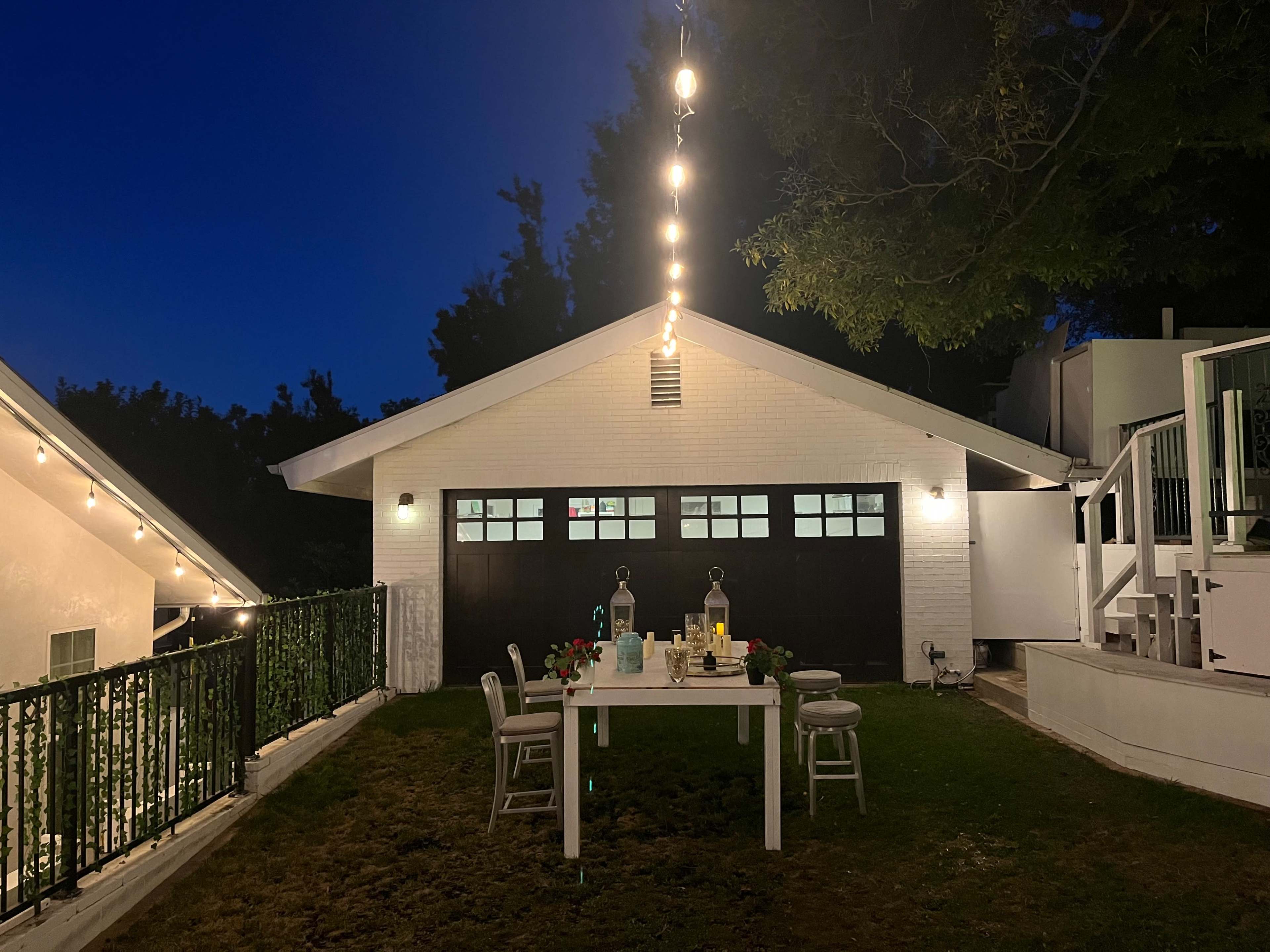 A table is set with beverages and flowers under string lights in a backyard near a white garage at night.