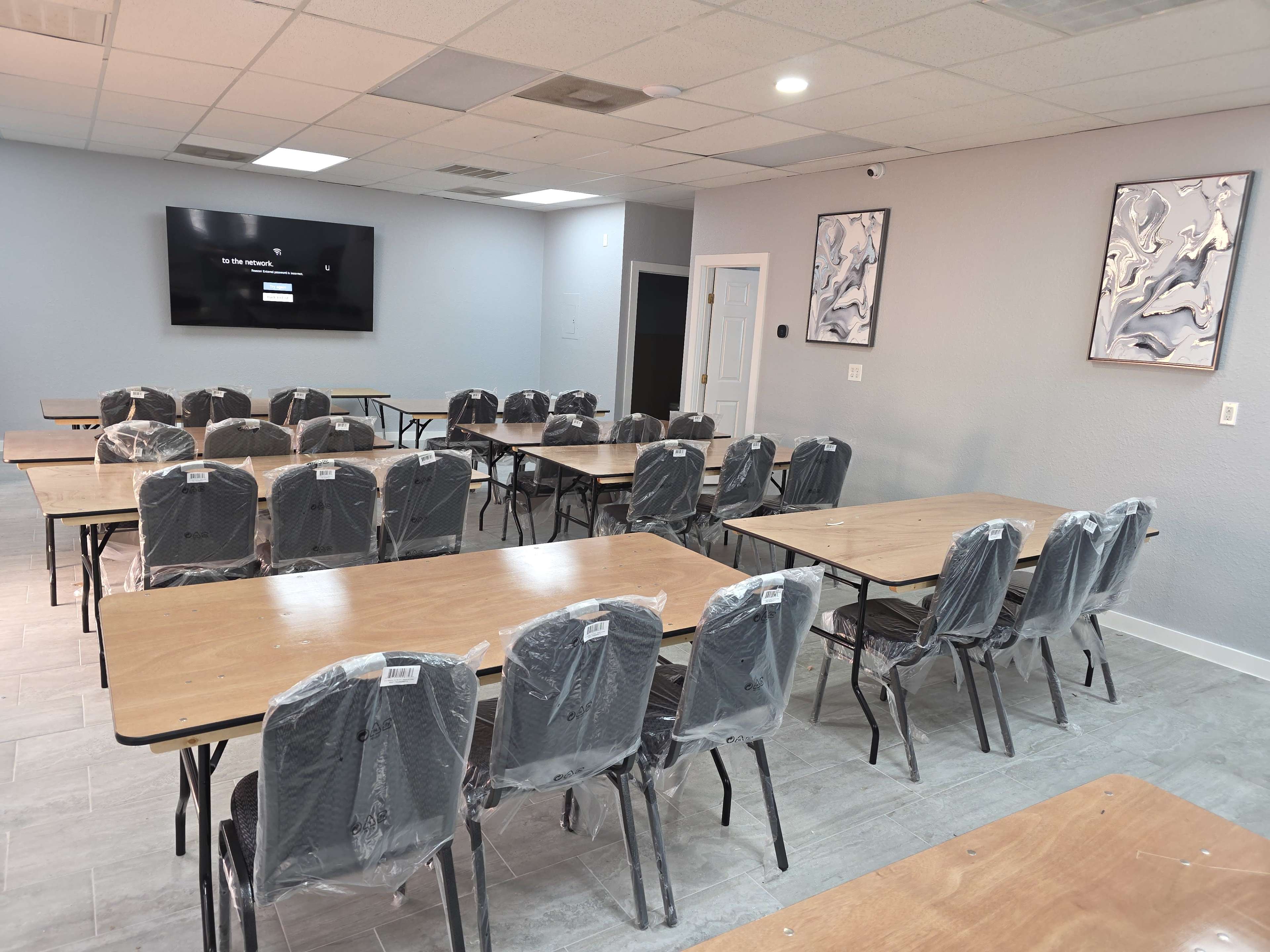 A conference room features multiple tables arranged in a grid pattern with plastic-covered chairs and a large screen on the wall.
