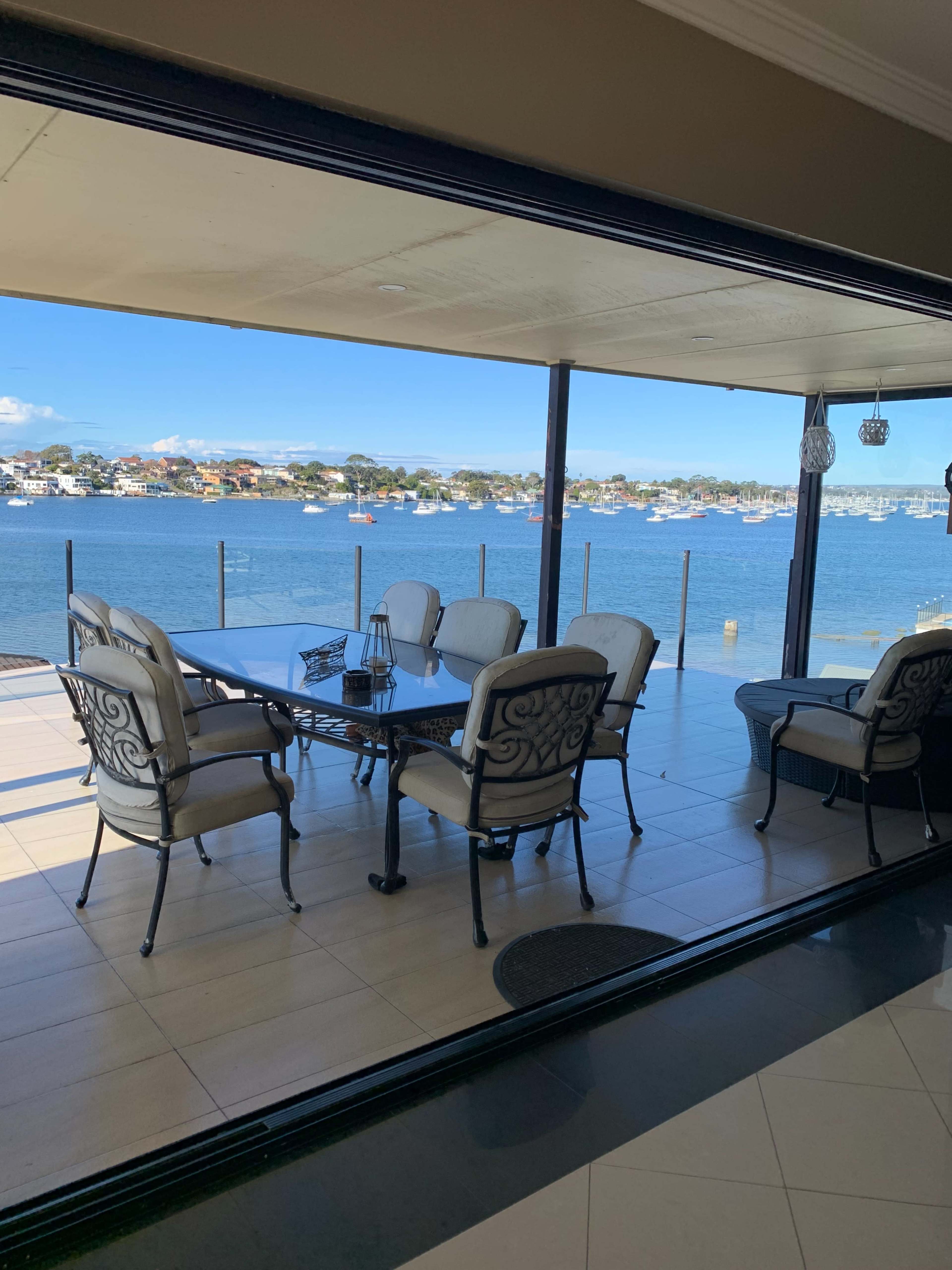 A covered patio with a table and chairs overlooks a waterfront view with boats and houses in the background.