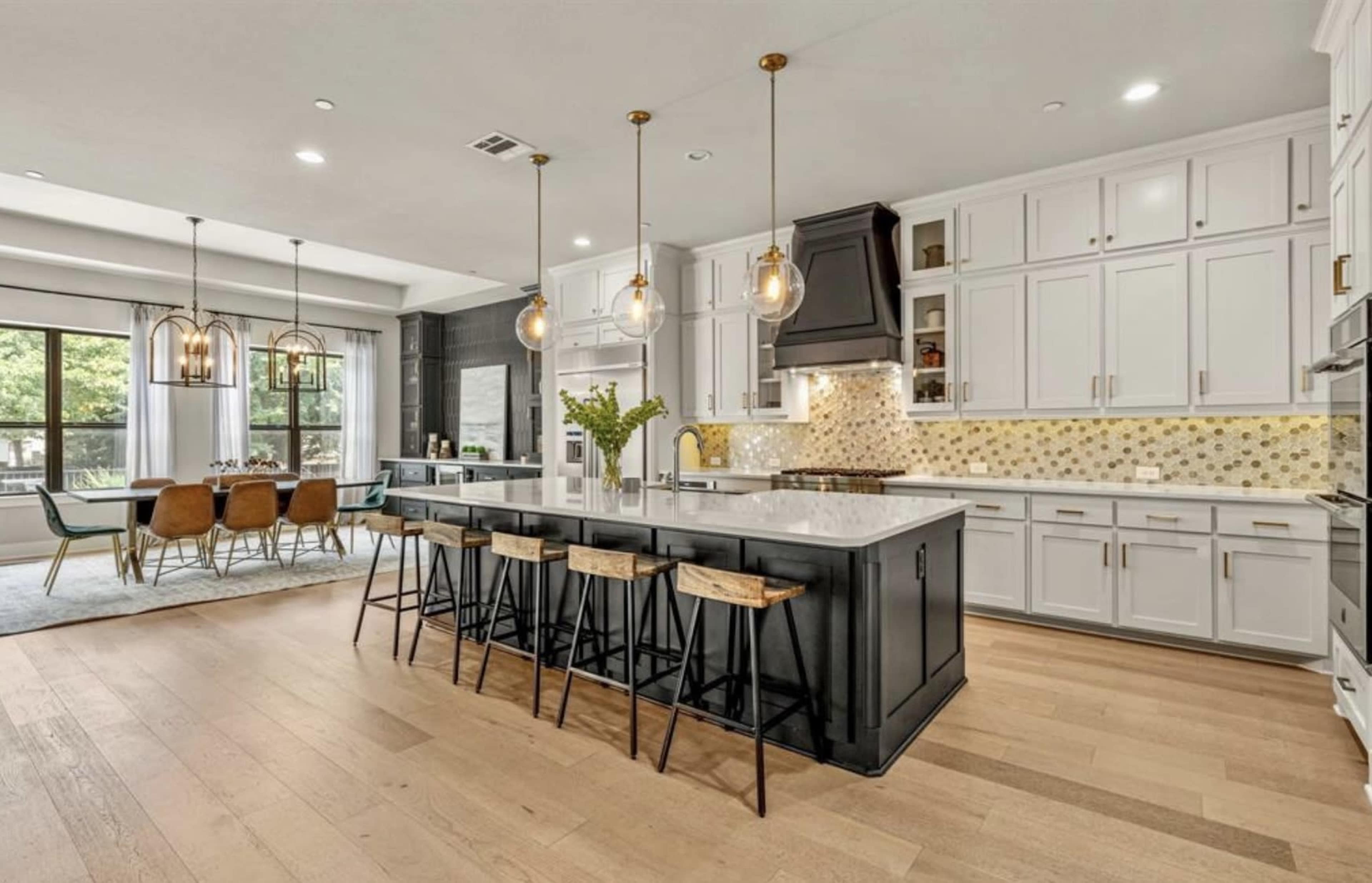 A modern kitchen features a large island with barstools, white cabinetry, and pendant lighting, alongside an adjacent dining area with a wooden table and chairs.