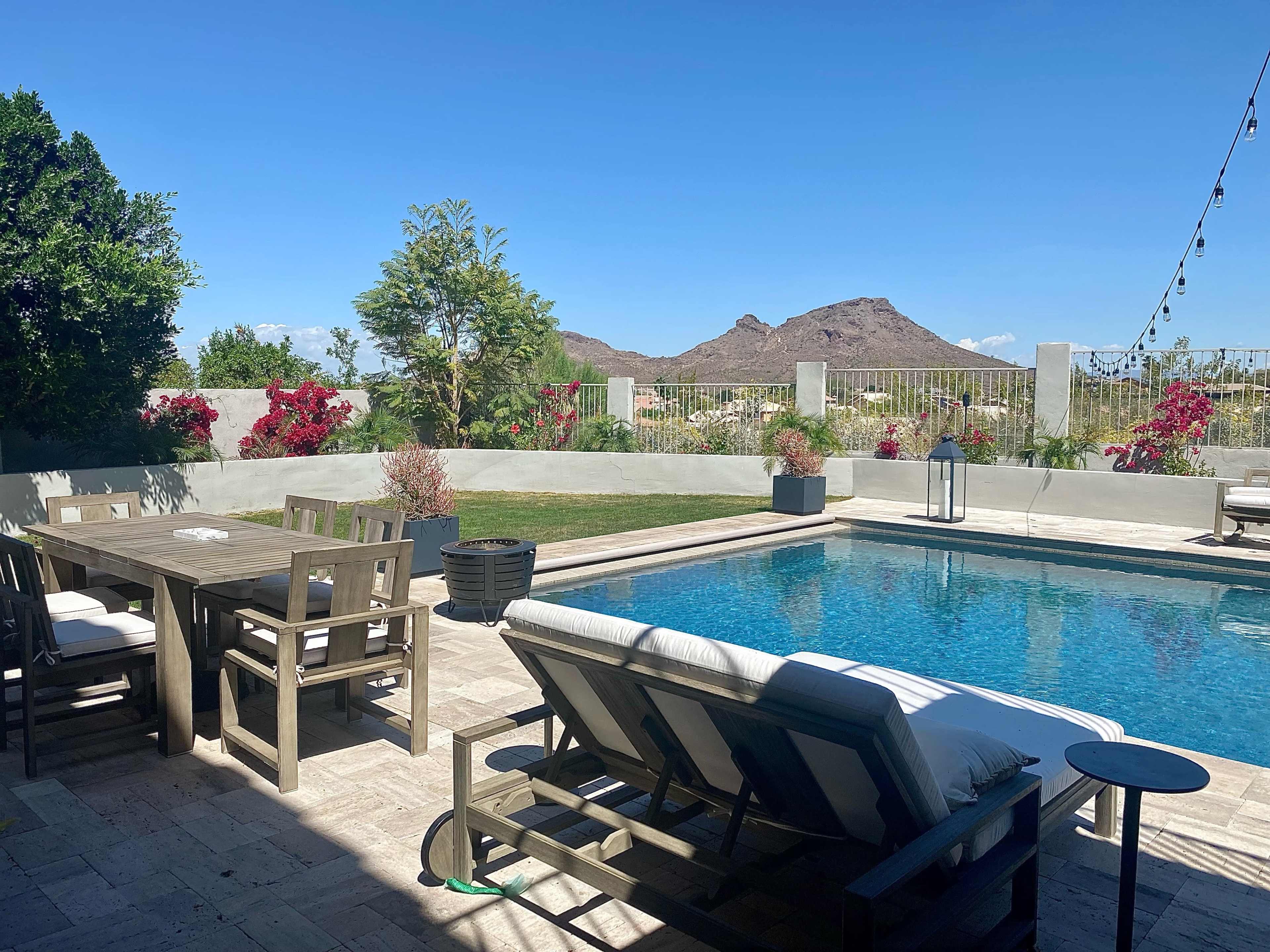 A patio area features a swimming pool surrounded by lounge chairs, a dining table, and colorful plants, set against a backdrop of mountains and a clear blue sky.