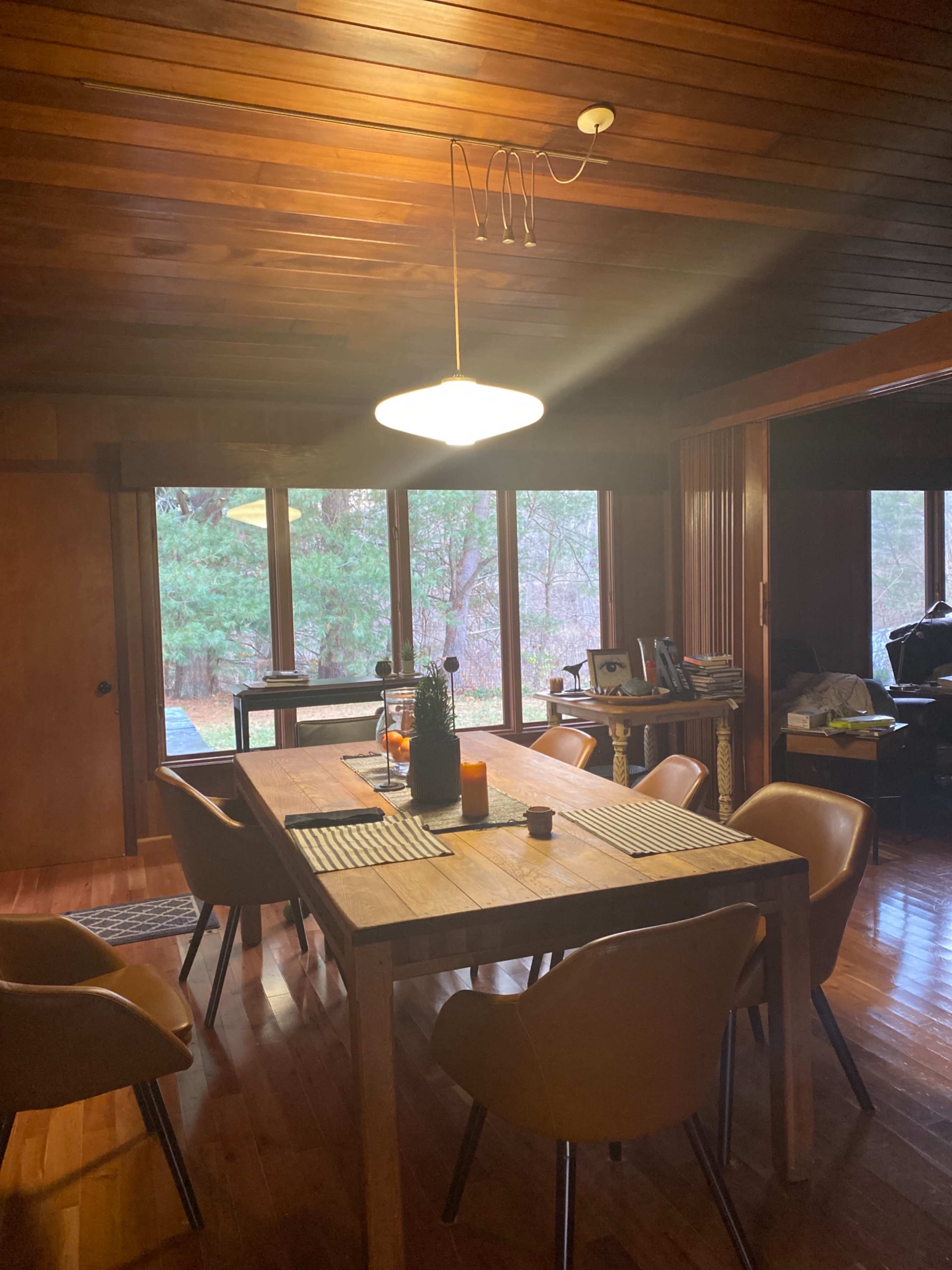 A wooden dining table is set with a table runner and chairs in a room with large windows overlooking a forested area.