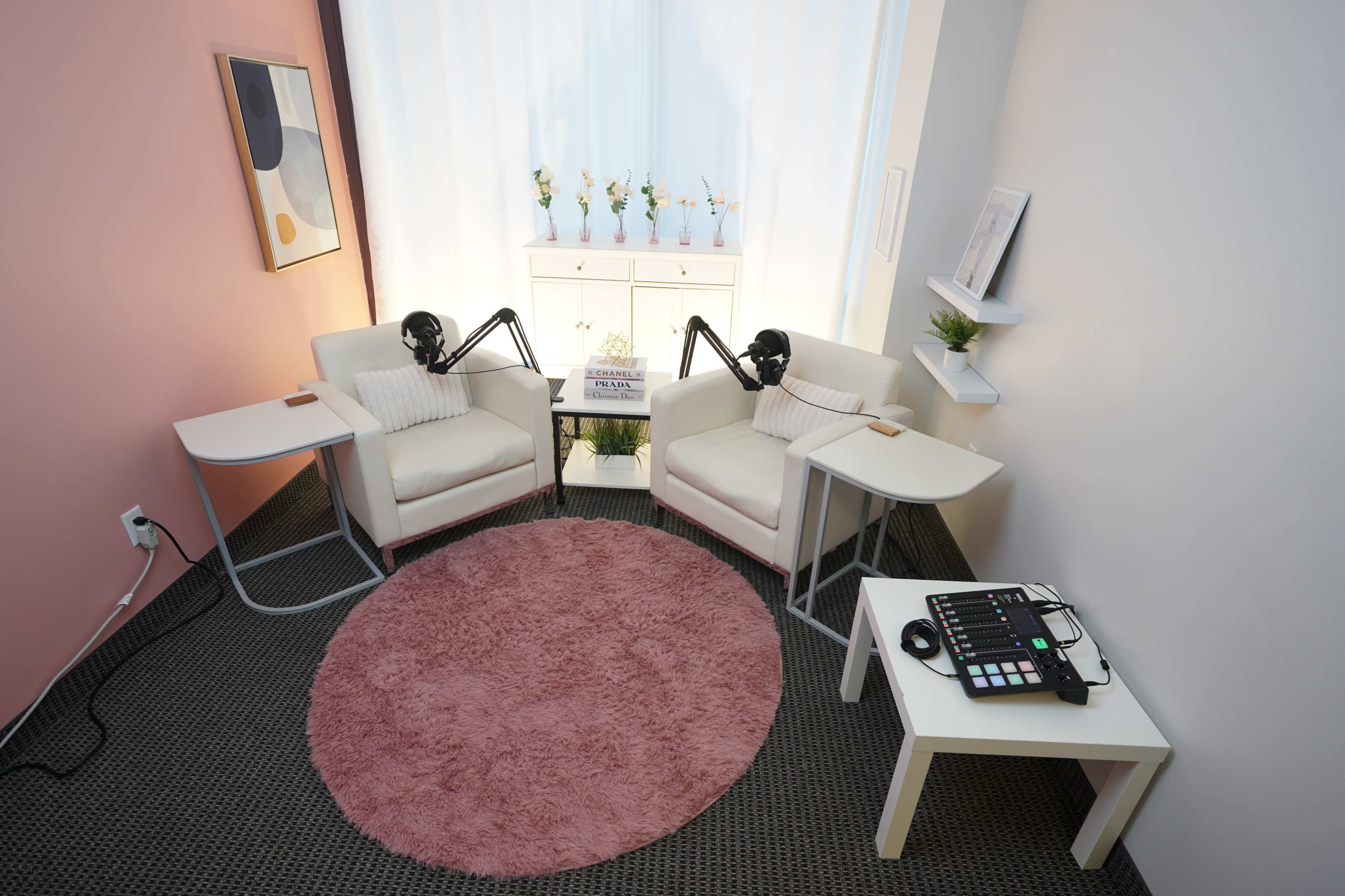 A cozy recording space features two white armchairs with black microphone stands, a round pink rug, and a small table with audio equipment.