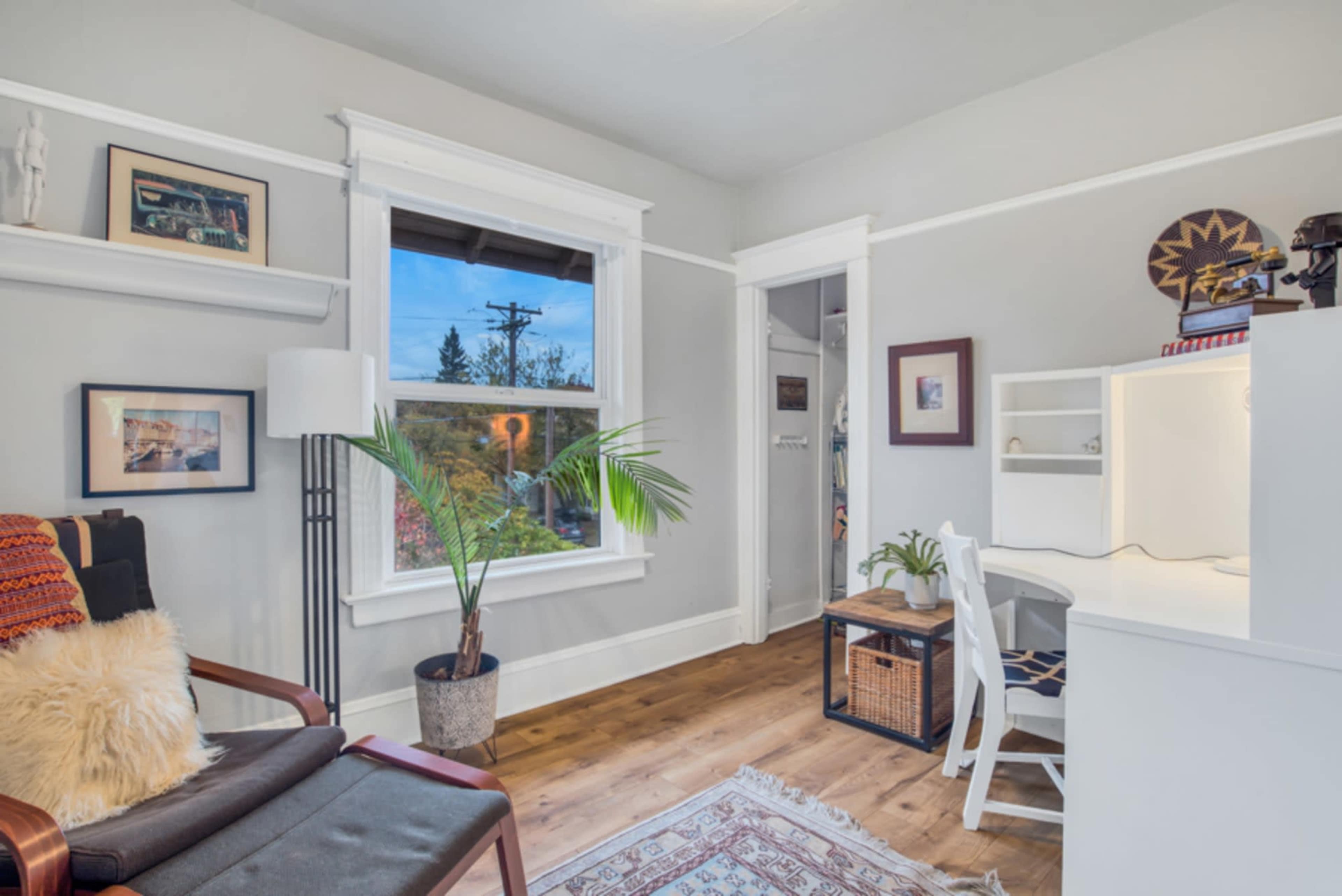 A cozy room features a desk with a chair, a plant in a pot, and a seating area with a comfortable chair and decorative throw, all against light gray walls.