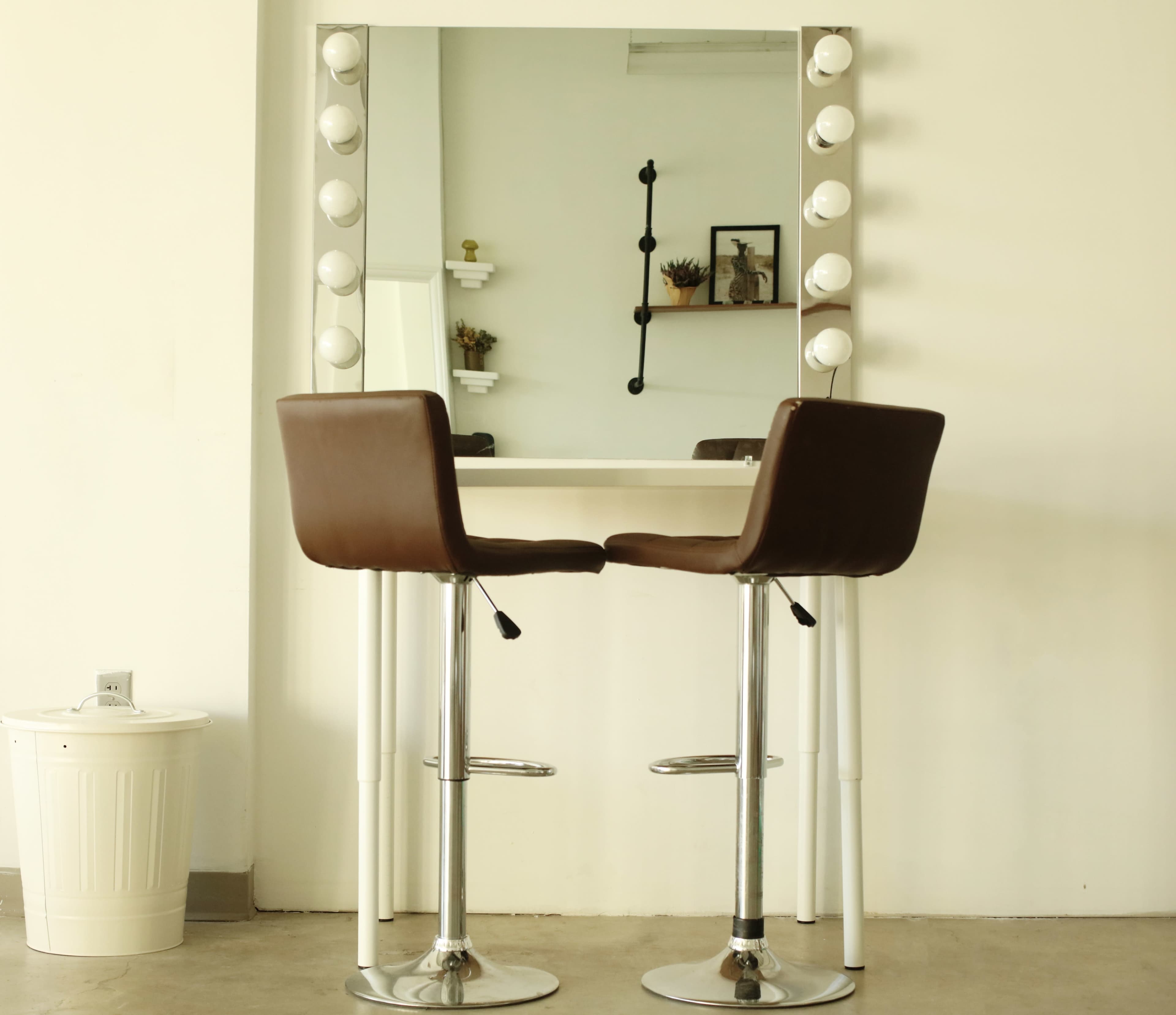 A minimalist dressing area features two brown bar stools in front of a large mirror framed by light bulbs, with a small decorative shelf in the background.