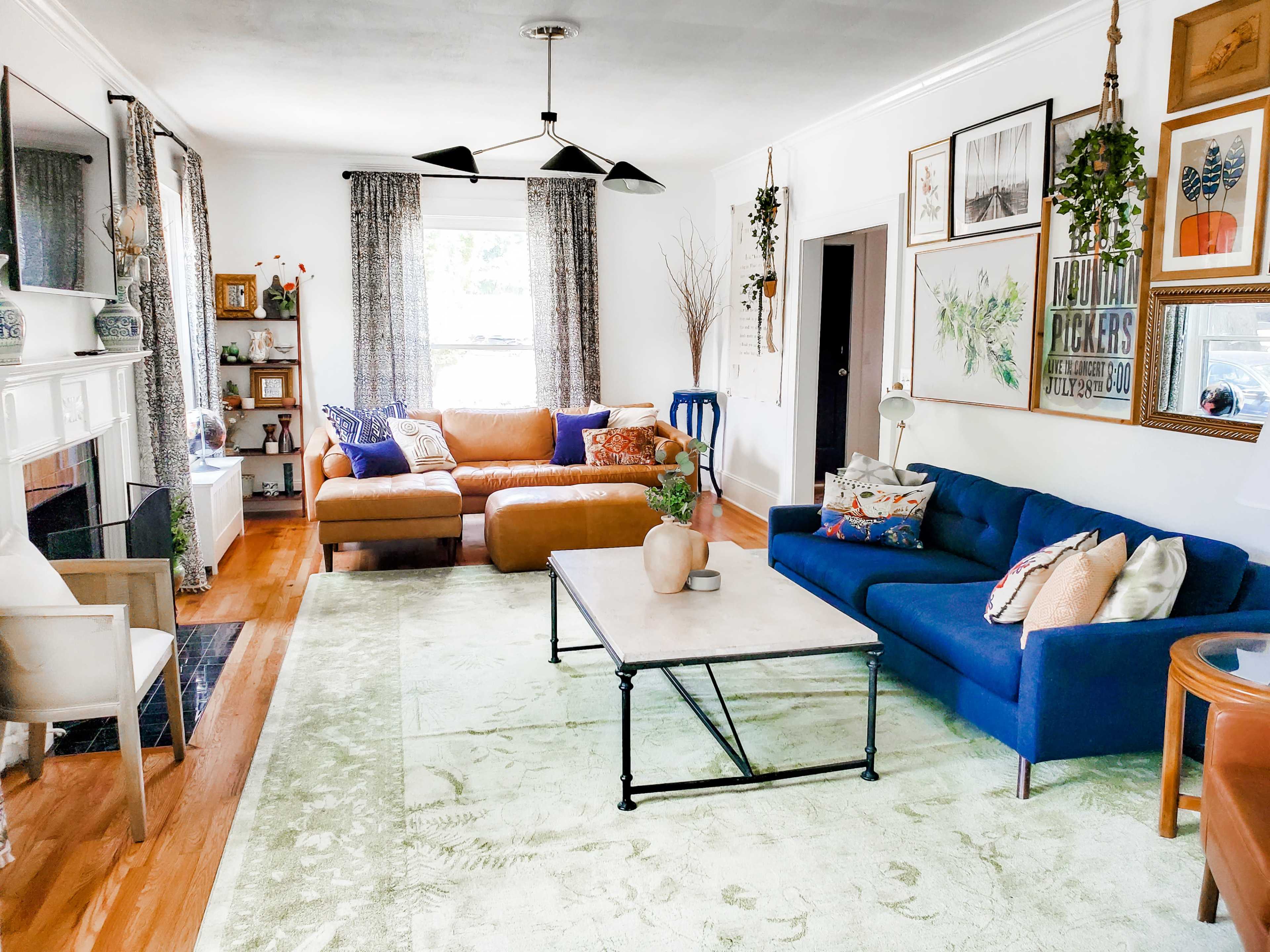 The living room features a blue sofa, a brown sectional, a coffee table, and various plants, framed artwork, and decorative items arranged around a light-colored rug.