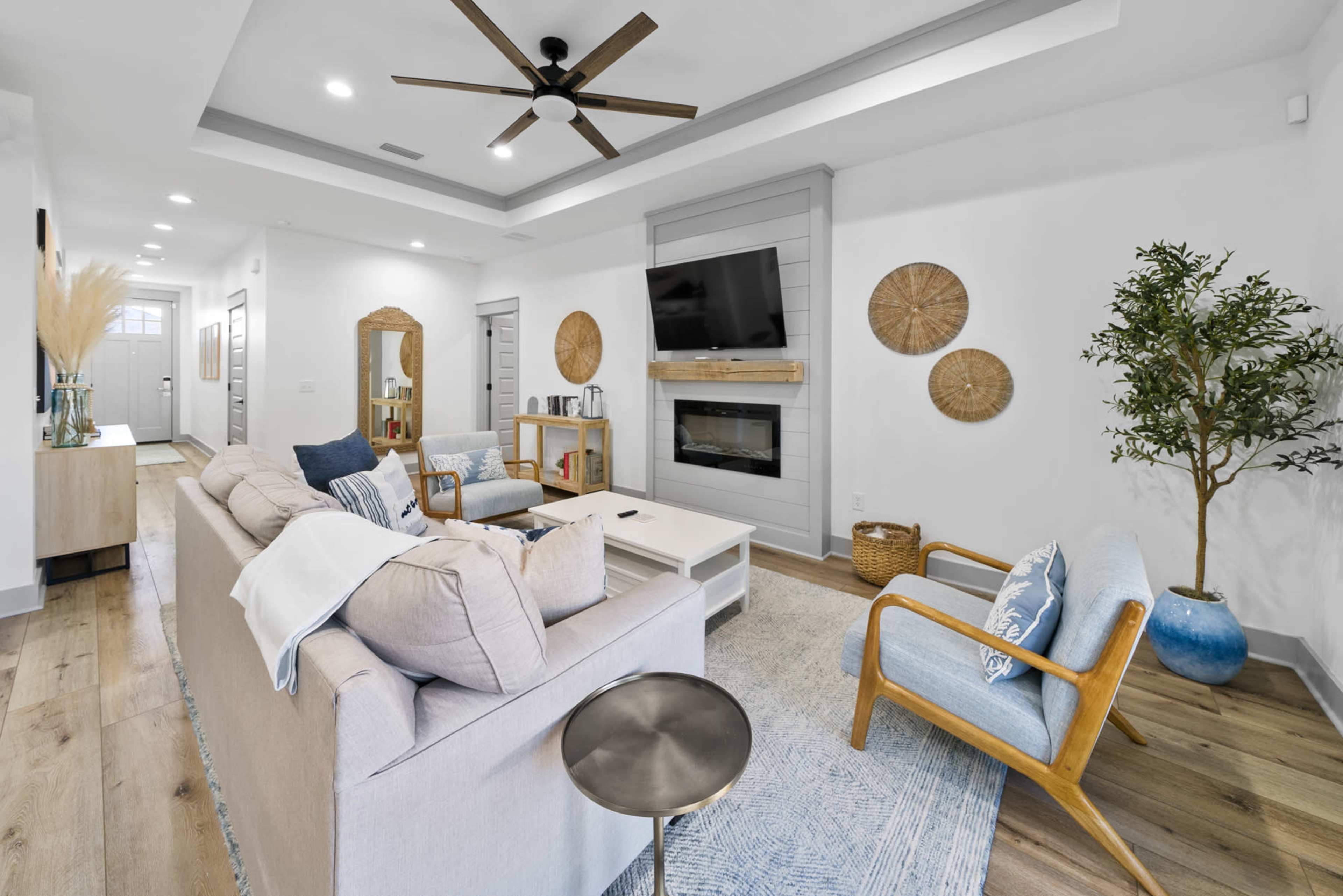 A modern living room with a light-colored sofa, a wooden coffee table, and a flat-screen TV mounted on a wall, complemented by decorative wall hangings and a plant.