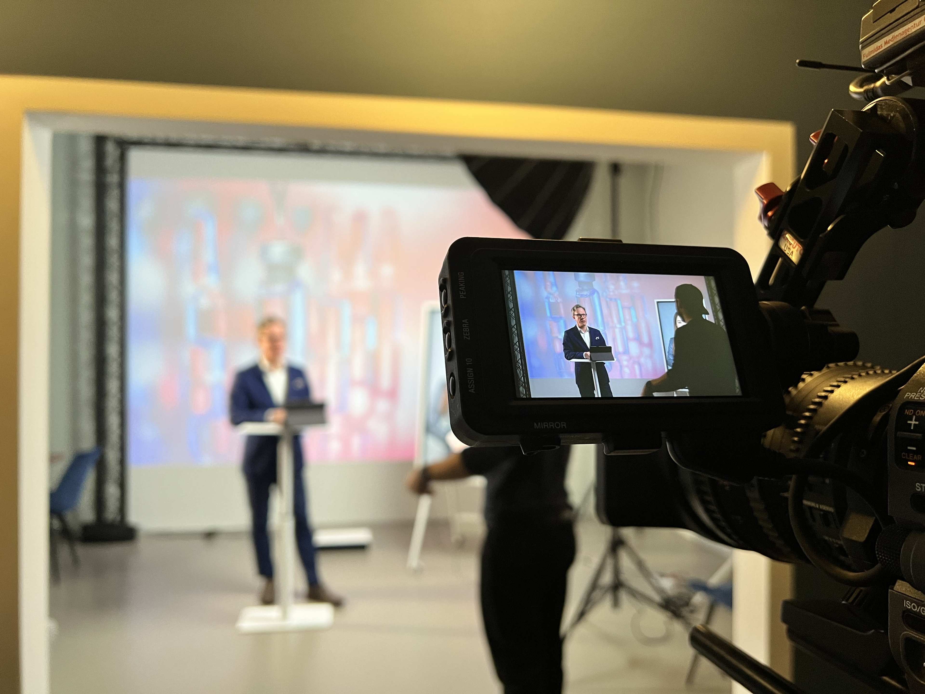 A speaker in a blue suit addresses an audience while standing behind a podium in a studio setting.