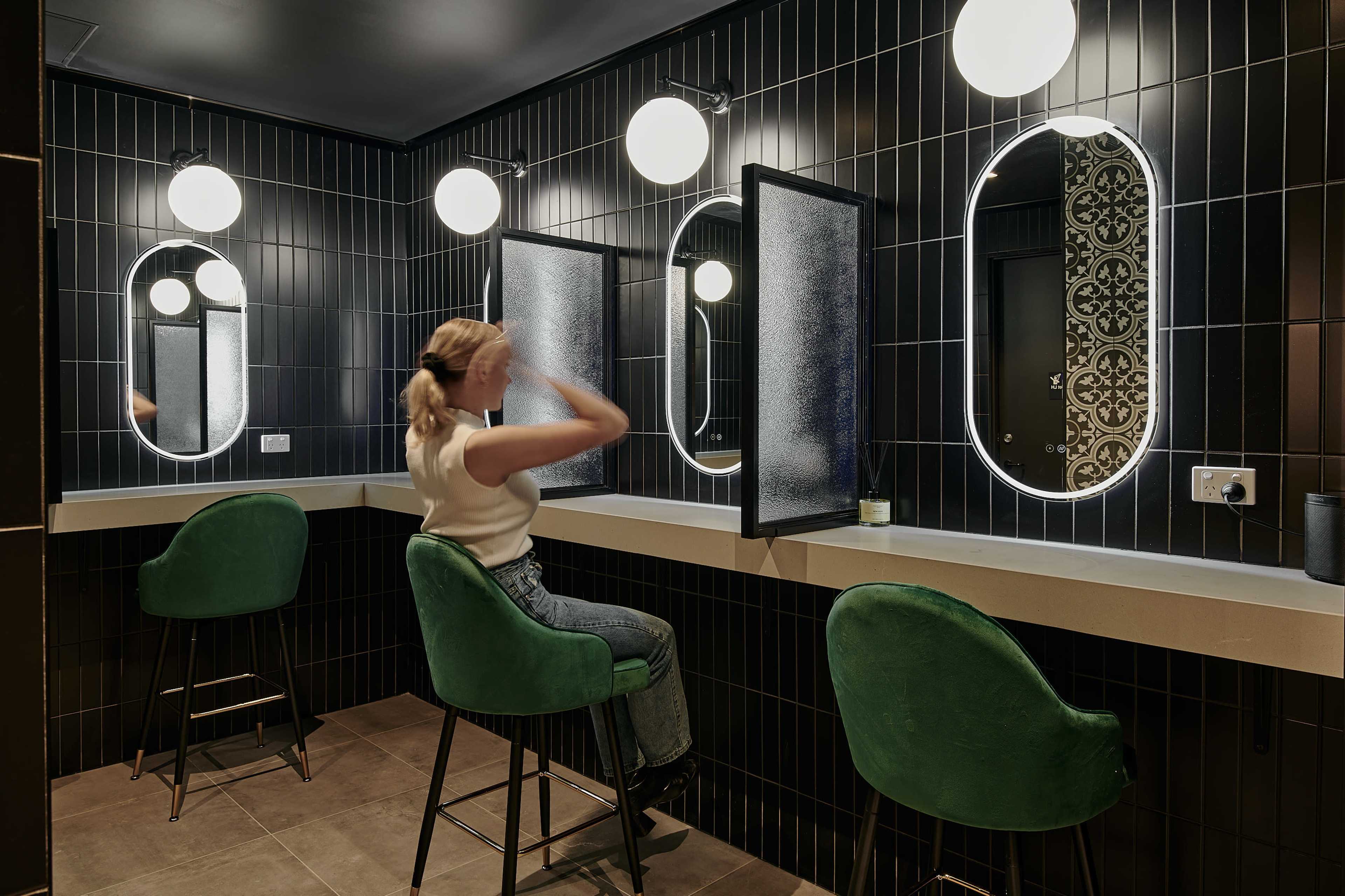 A person sits at a countertop in a dimly lit bathroom with black tiled walls and illuminated mirrors reflecting a stylish modern design.