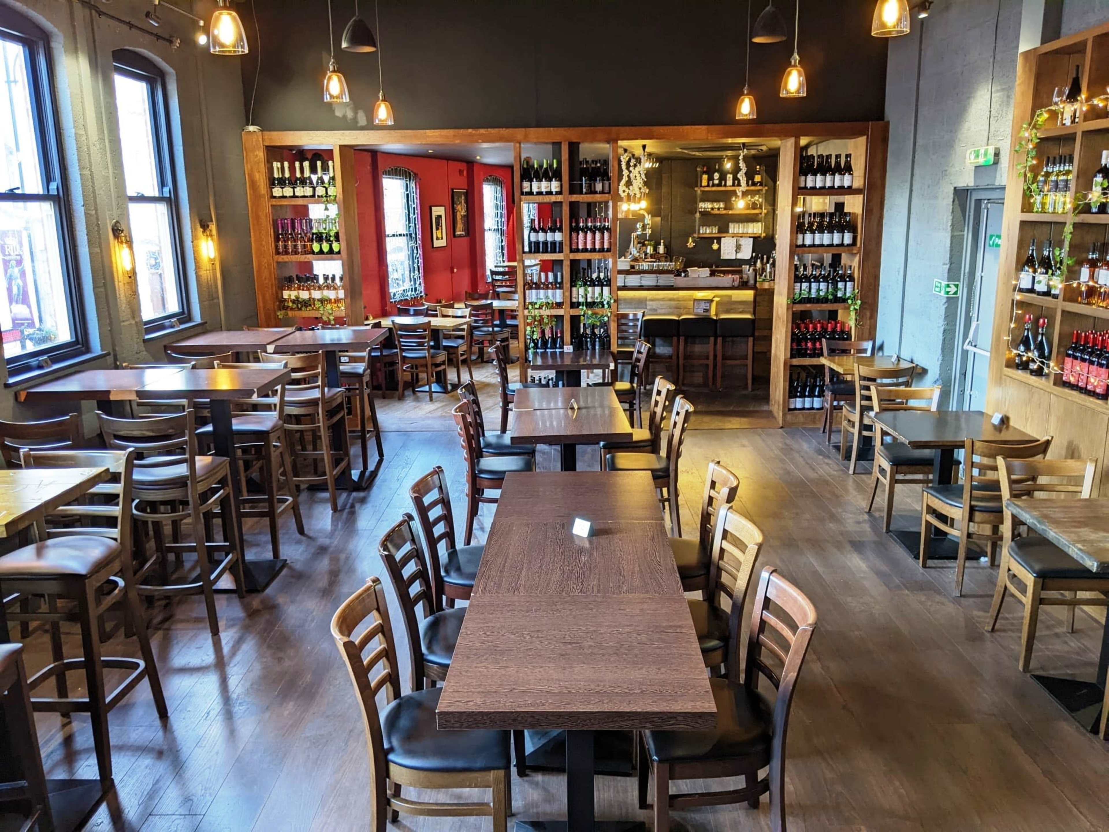 A spacious restaurant interior with wooden tables and chairs arranged around a central bar, flanked by shelves filled with bottles.