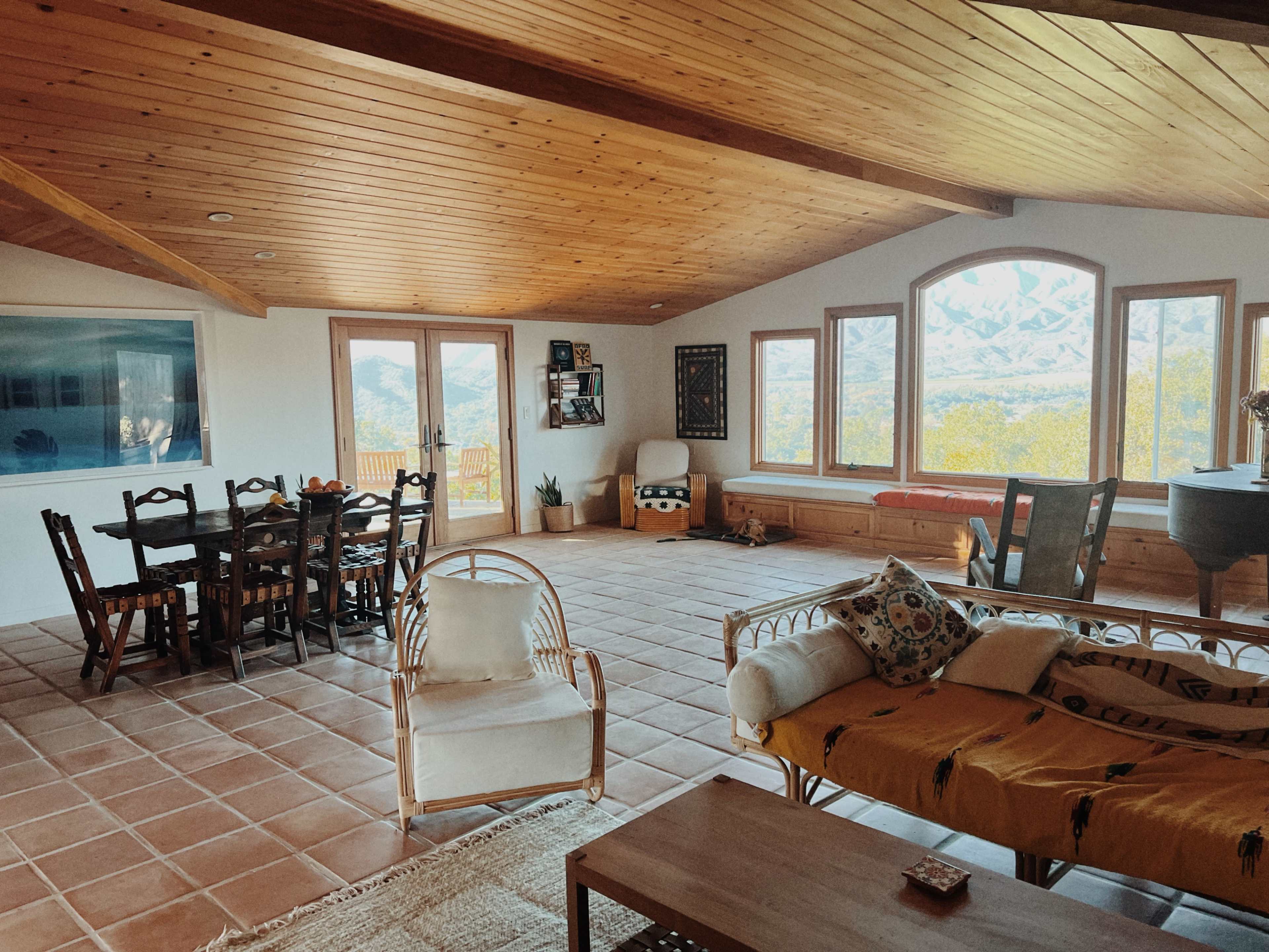 A spacious living area features a wooden ceiling, large windows with mountain views, a dining table, and comfortable seating arrangements.