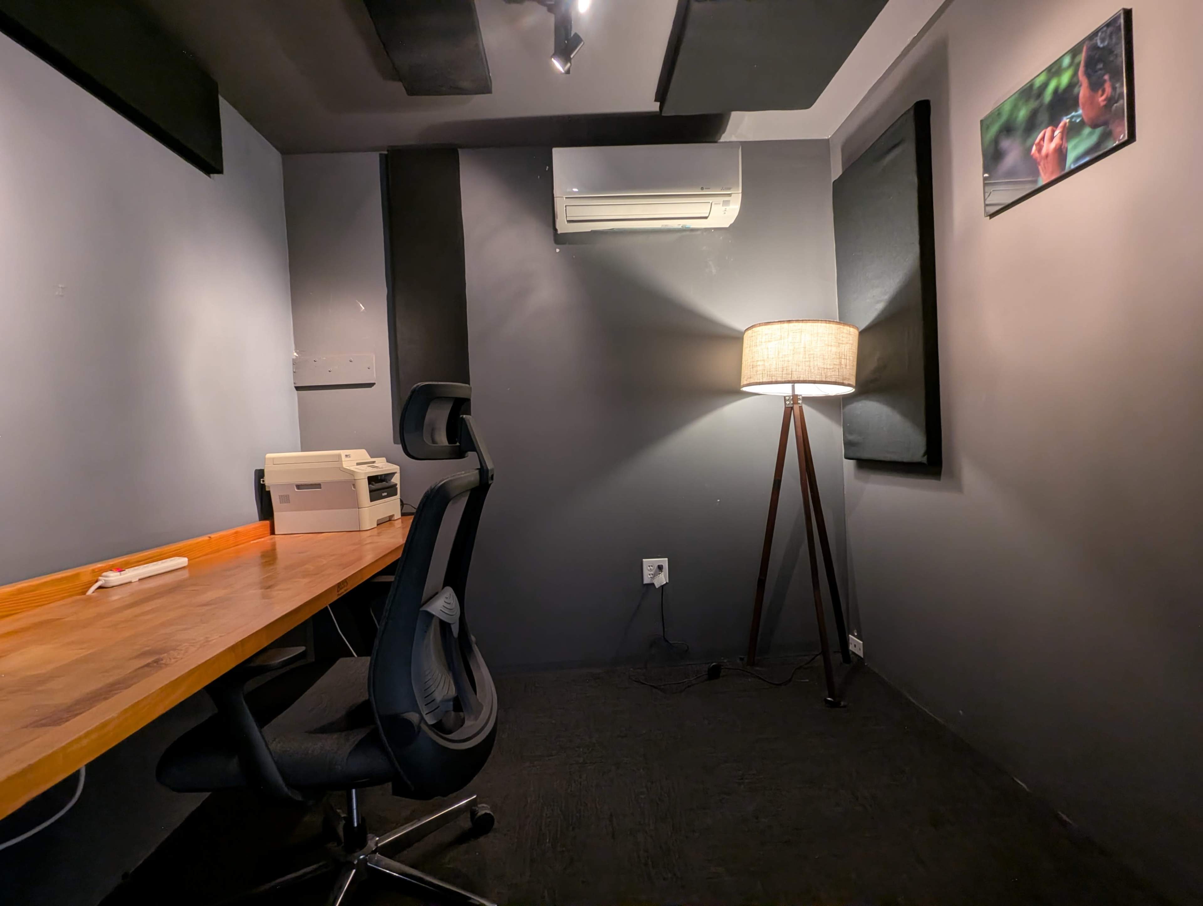 The image shows a small, sparse office space featuring a wooden desk with a chair, a lamp on a stand, and an air conditioning unit mounted on the wall.