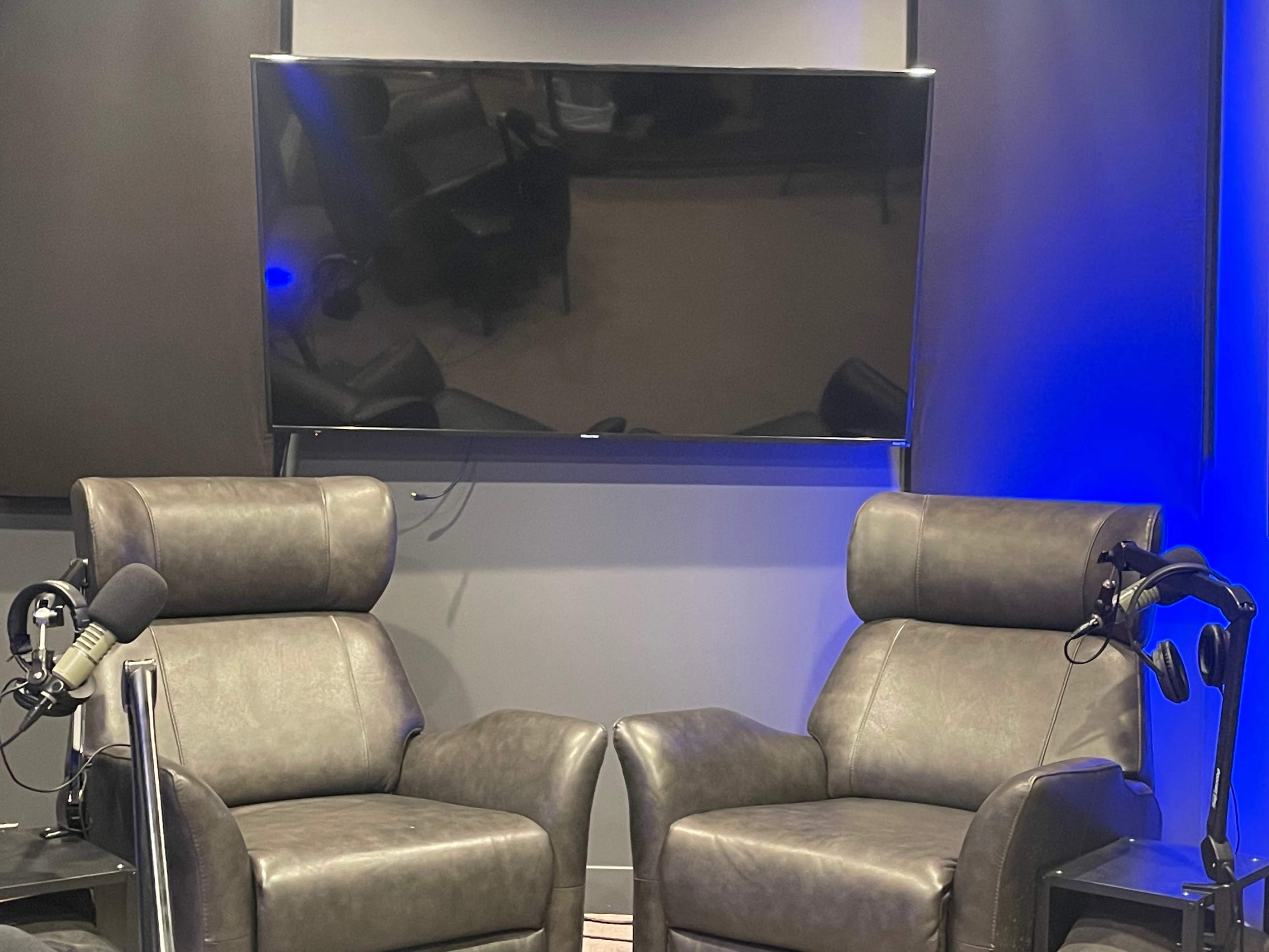 Two leather chairs facing each other in a recording studio, with a large flat-screen TV mounted on the wall behind them.