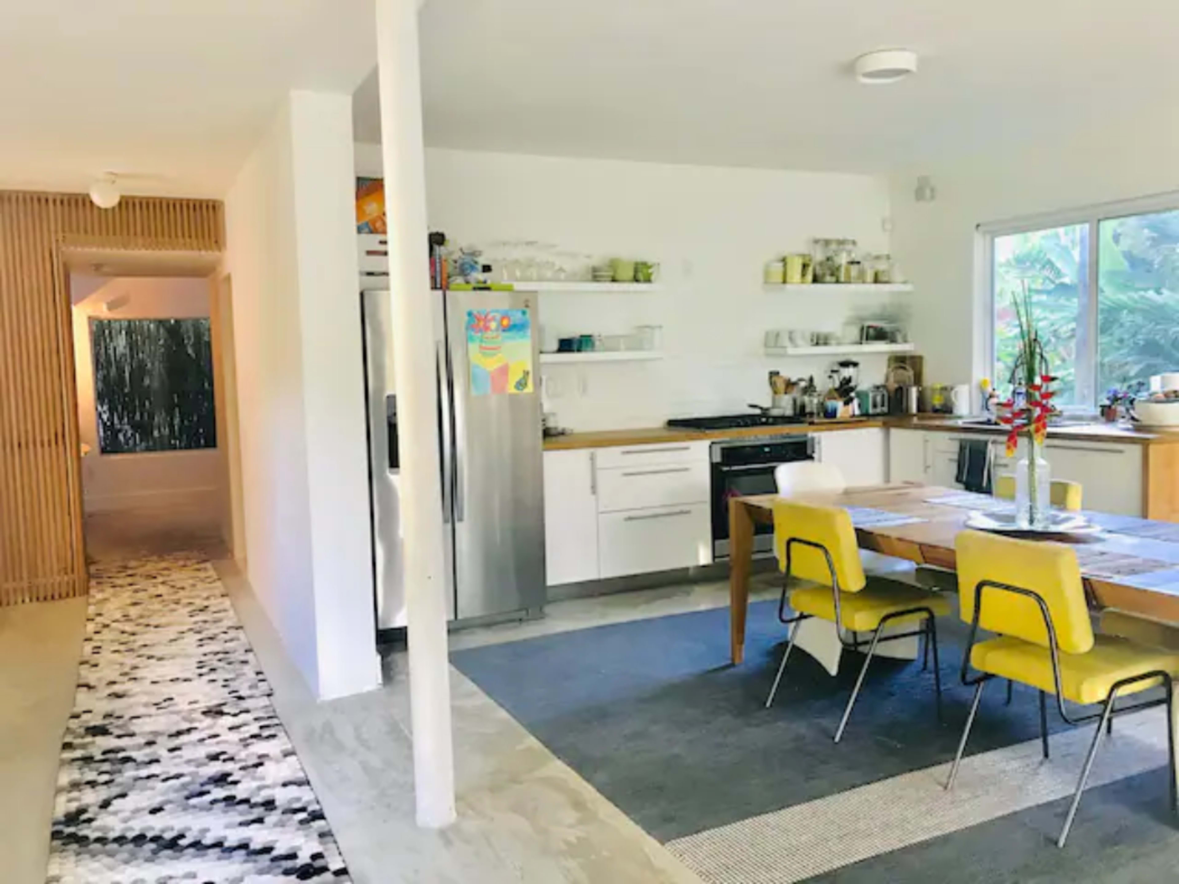 A modern kitchen and dining area with a wooden table, yellow chairs, and stainless steel appliances, leading to a hallway with a patterned runner.