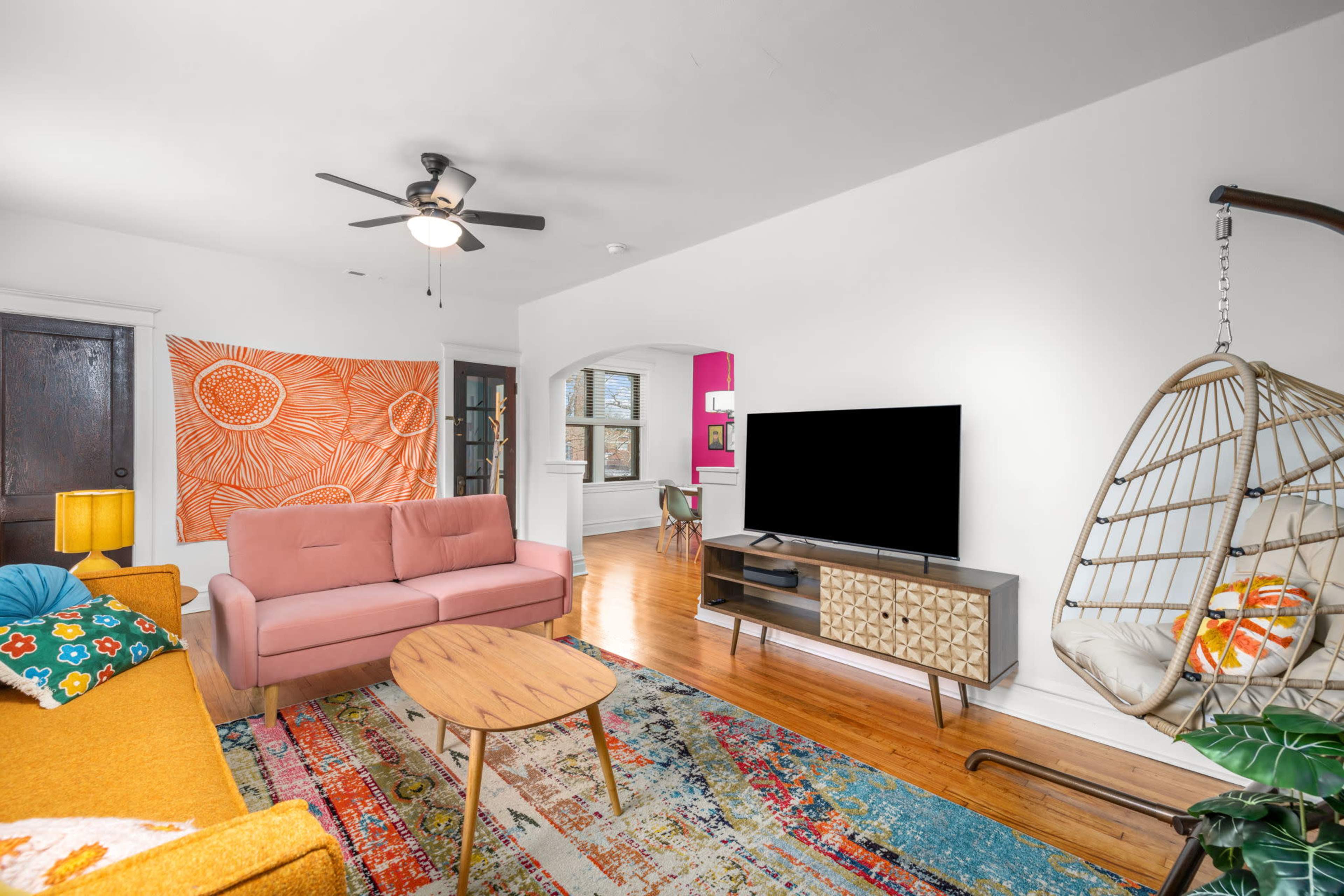 The living room features a pink sofa, a yellow armchair, a patterned rug, and a hanging chair, with a large TV and vibrant wall art.