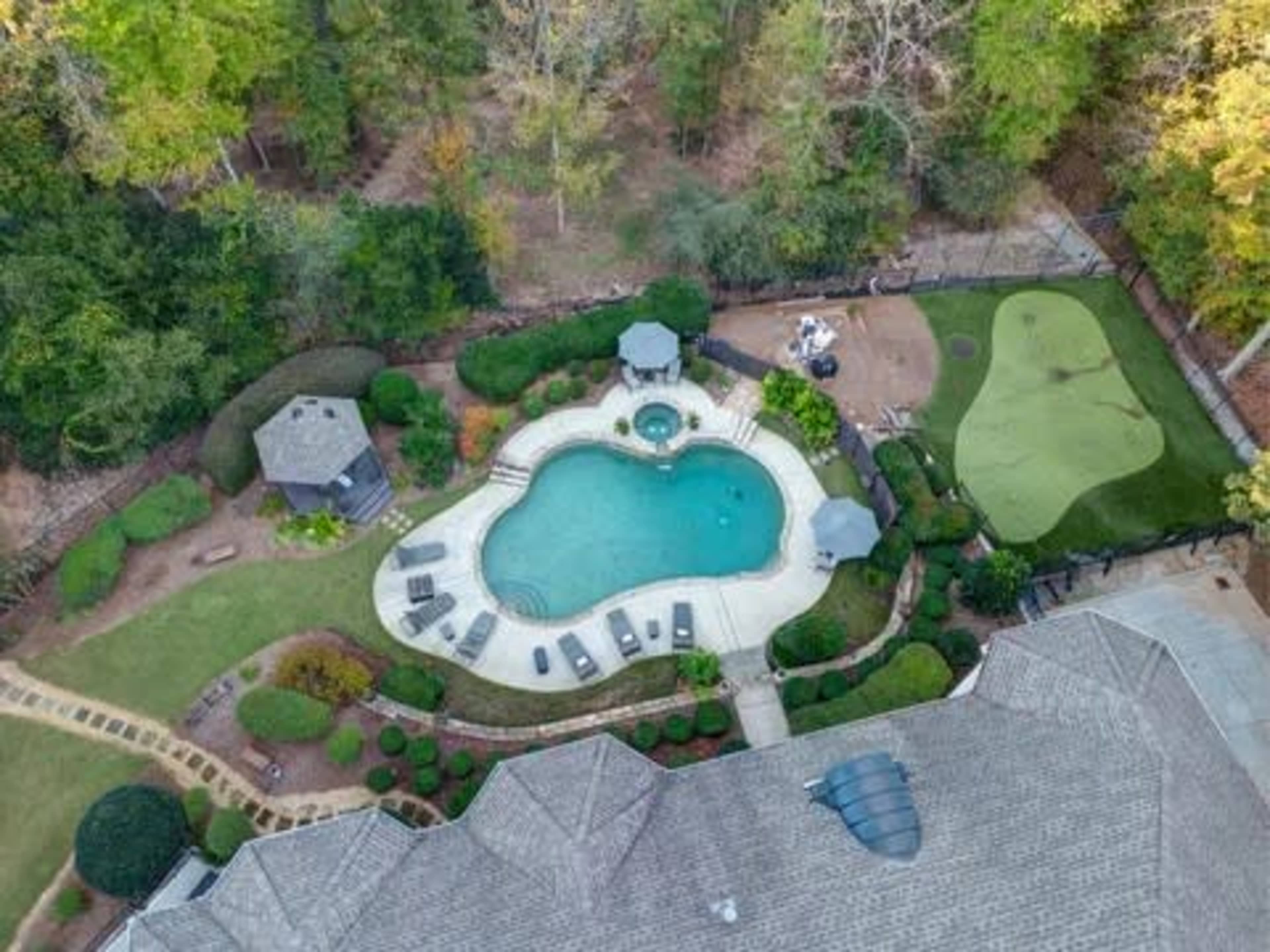 The image shows an aerial view of a backyard featuring a swimming pool, hot tub, and a putting green surrounded by greenery and landscaped areas.