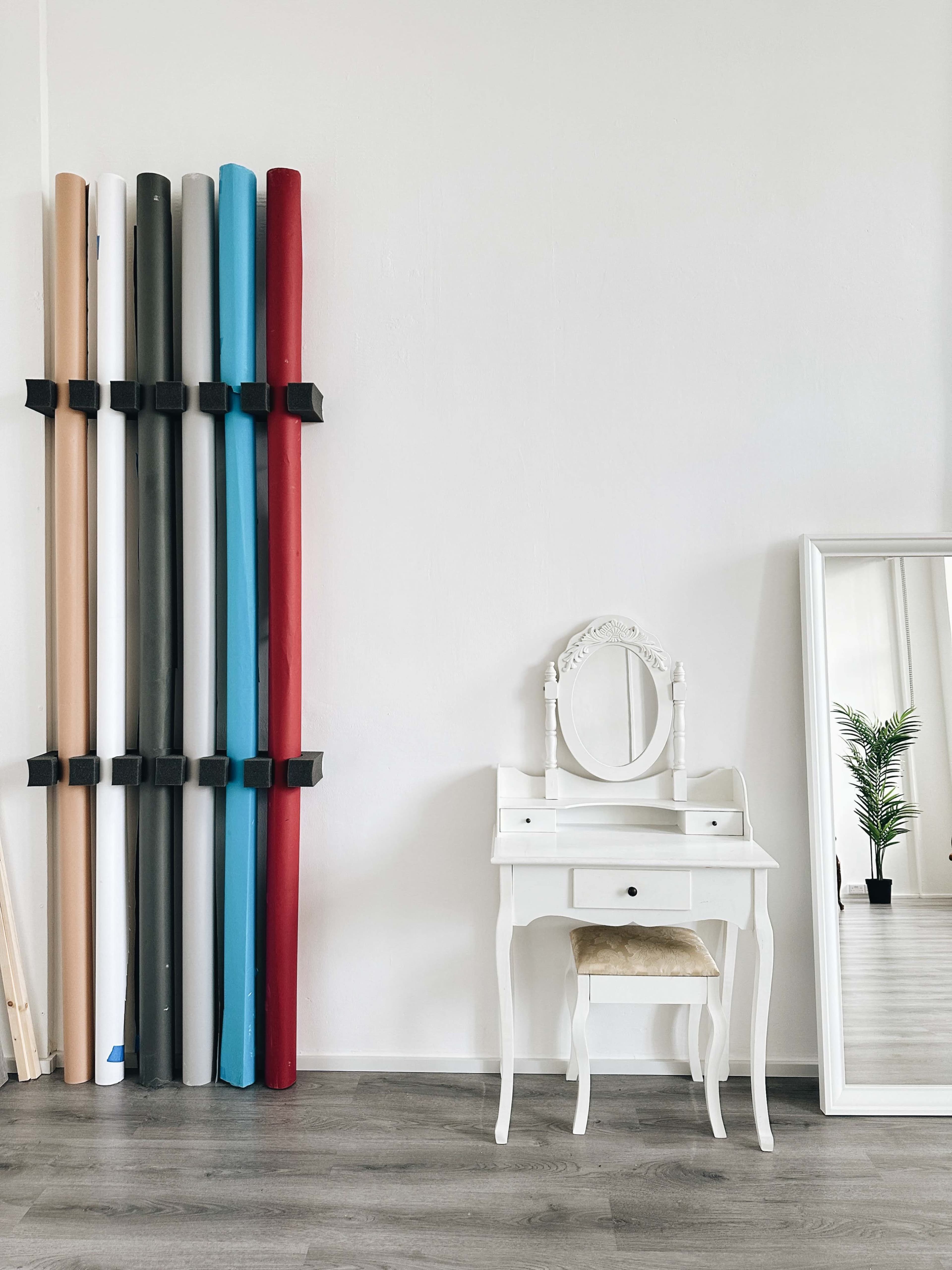 The image shows a simple, well-lit room featuring a stack of colored backdrop rolls secured to the wall and a white vanity table with an ornate mirror positioned next to it.