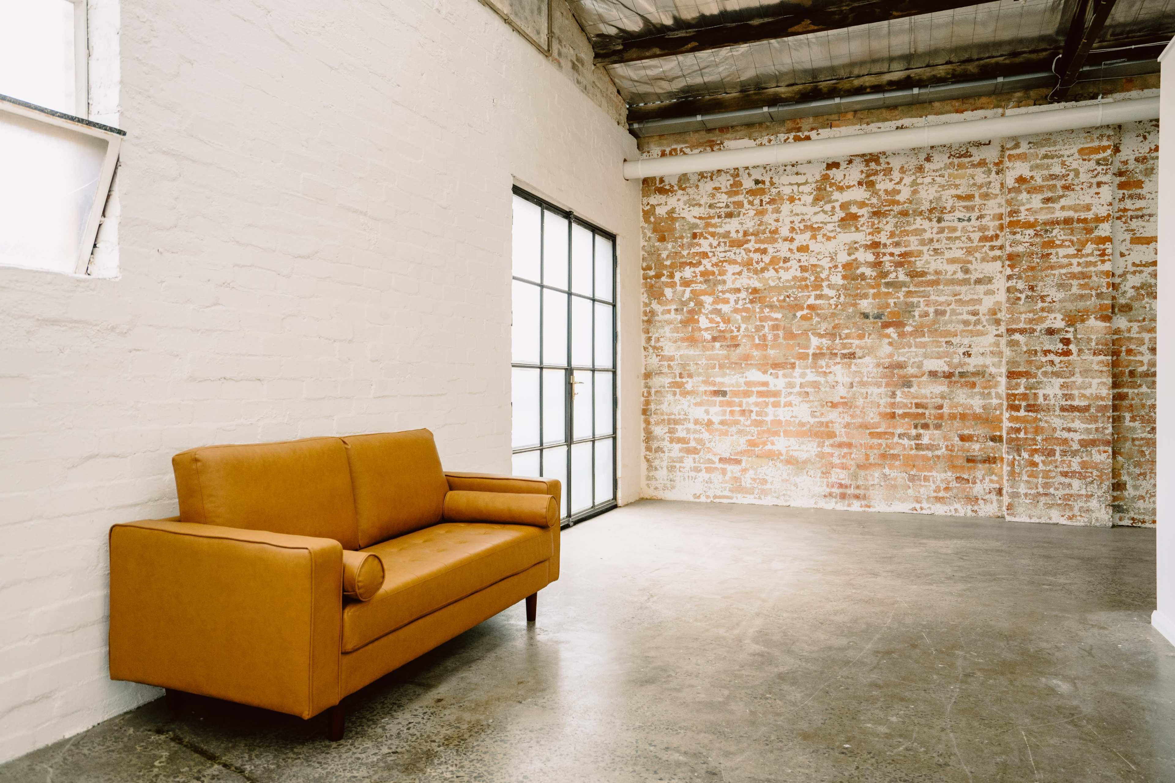 A rust-colored sofa sits against a brick wall in a spacious, industrial-style interior with large windows.