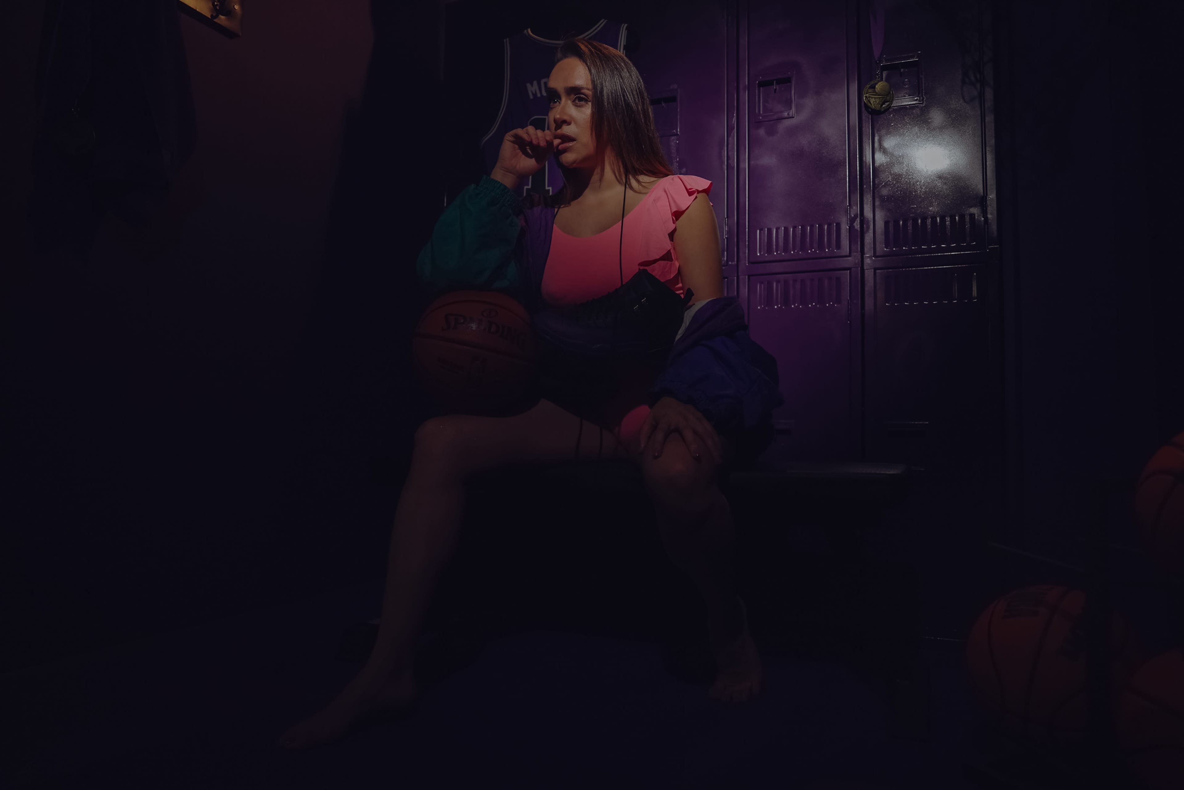 A woman in a pink outfit and colorful jacket sits on a bench in front of purple lockers, holding a basketball.