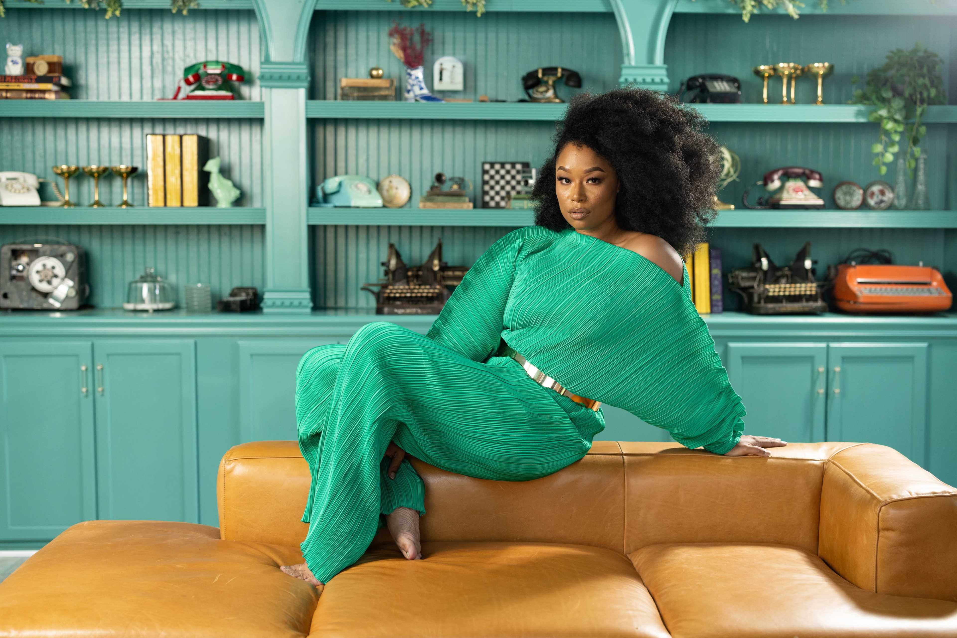 A woman in a green pleated outfit sits on a tan leather couch in front of a teal shelving unit filled with vintage items.